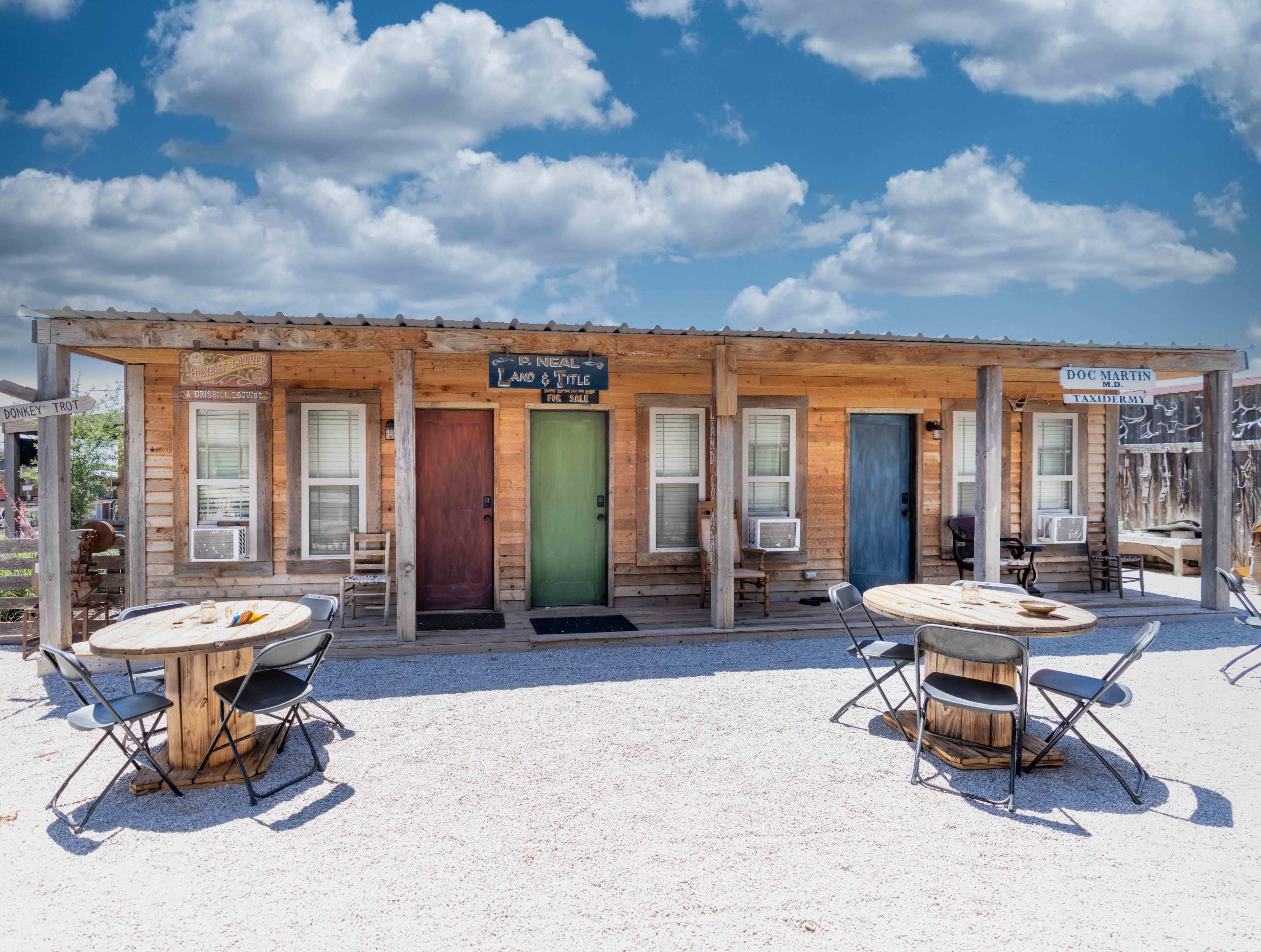 The image shows a row of small rustic wooden cabins with colorful doors and outdoor tables in a gravel area under a blue sky with clouds.
