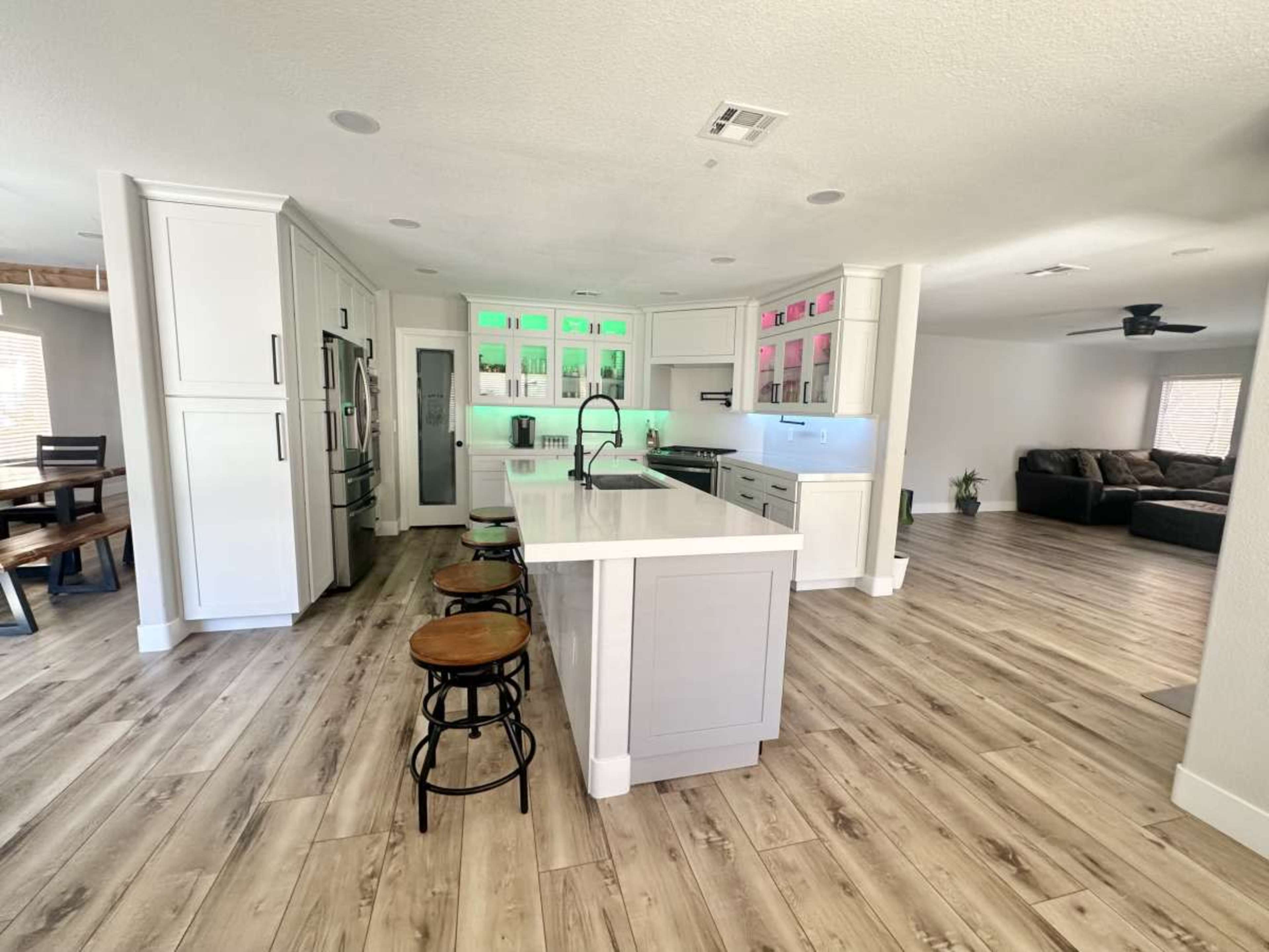 A modern kitchen features a central island with bar stools, sleek cabinetry, and colorful lighting behind the glass shelving.