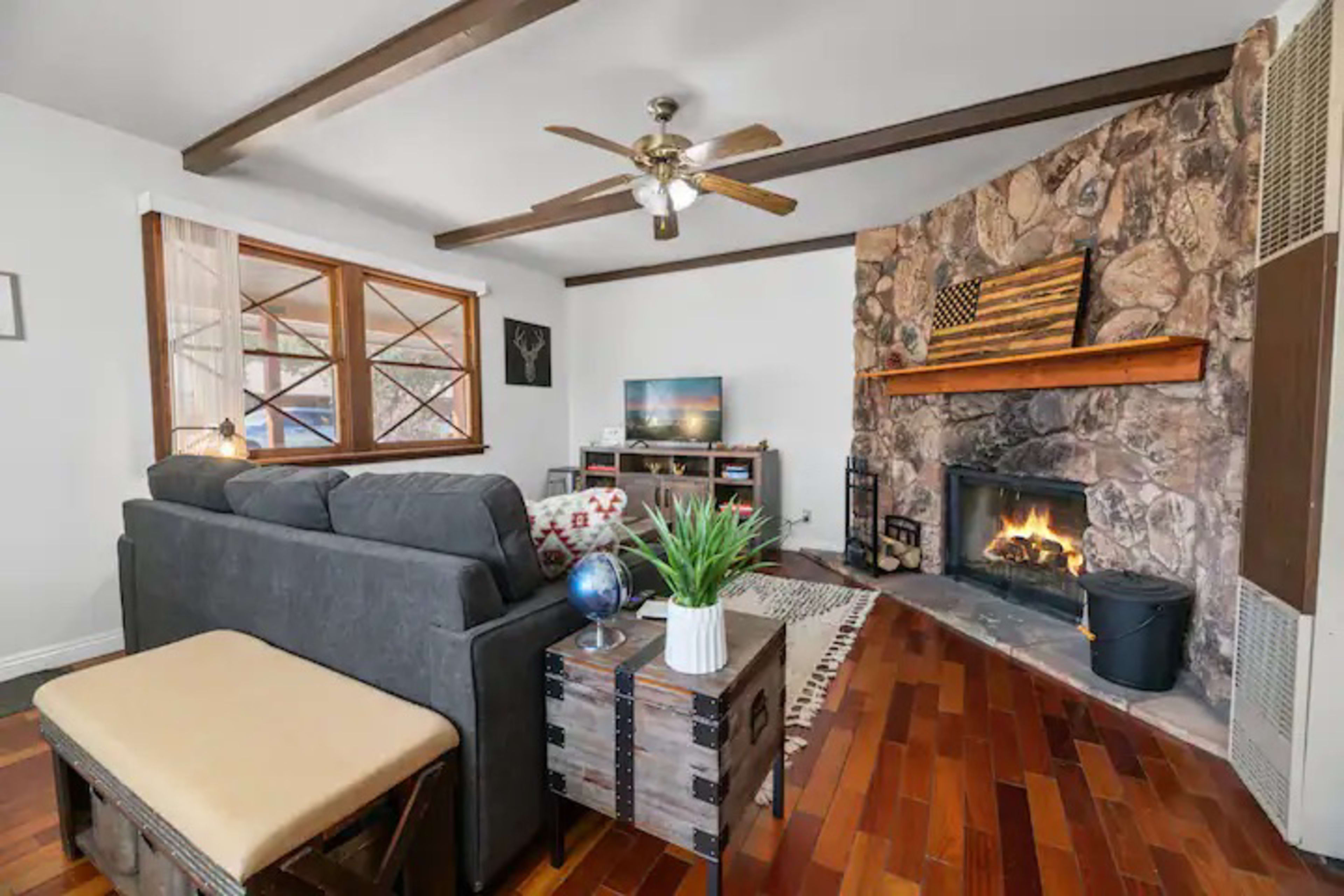 A living room with a stone fireplace, a gray sofa, wooden flooring, and a television on a stand near a window.