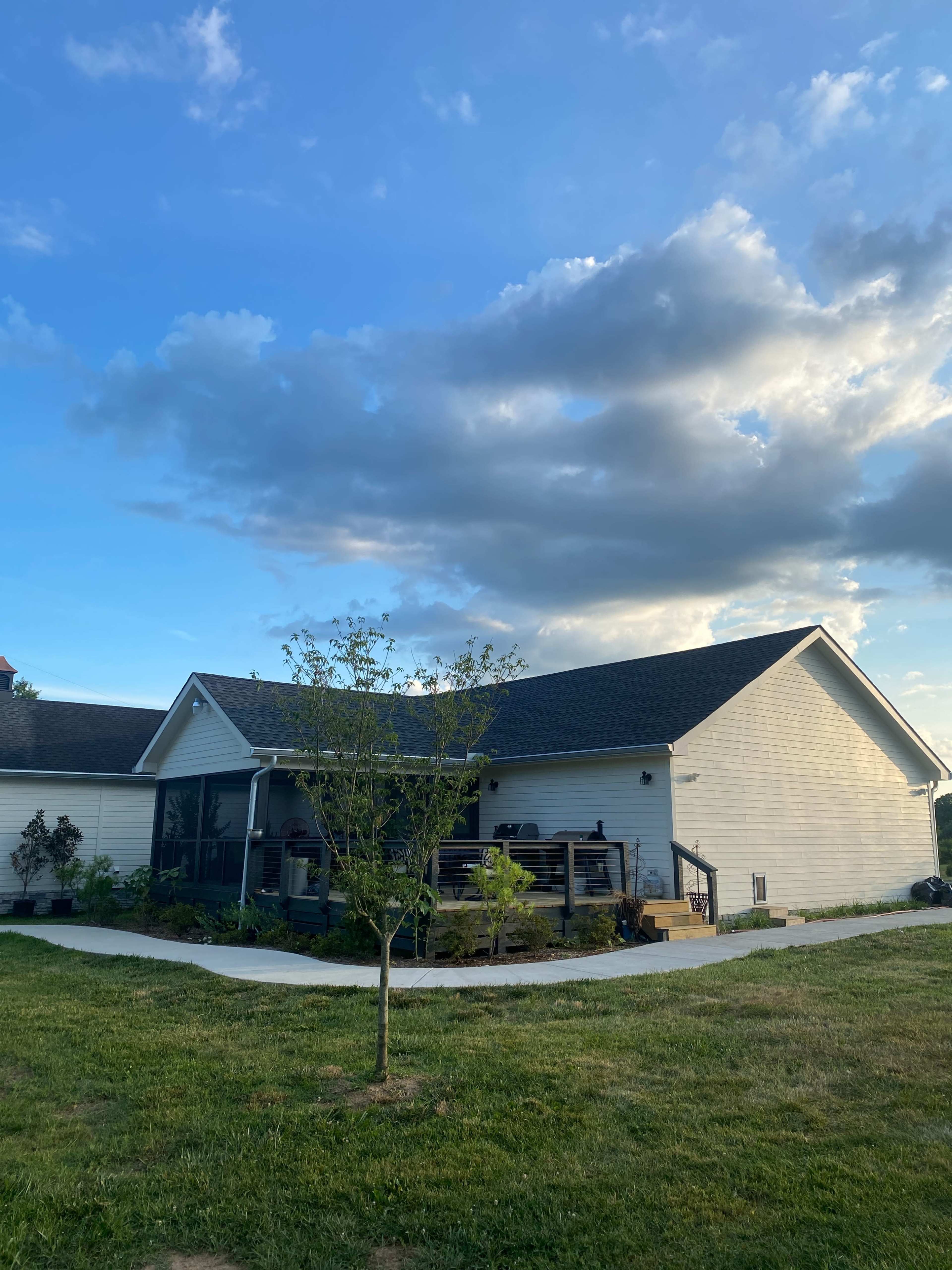 A single-story house with a patio surrounded by green grass and a small tree nearby, set against a partly cloudy sky.