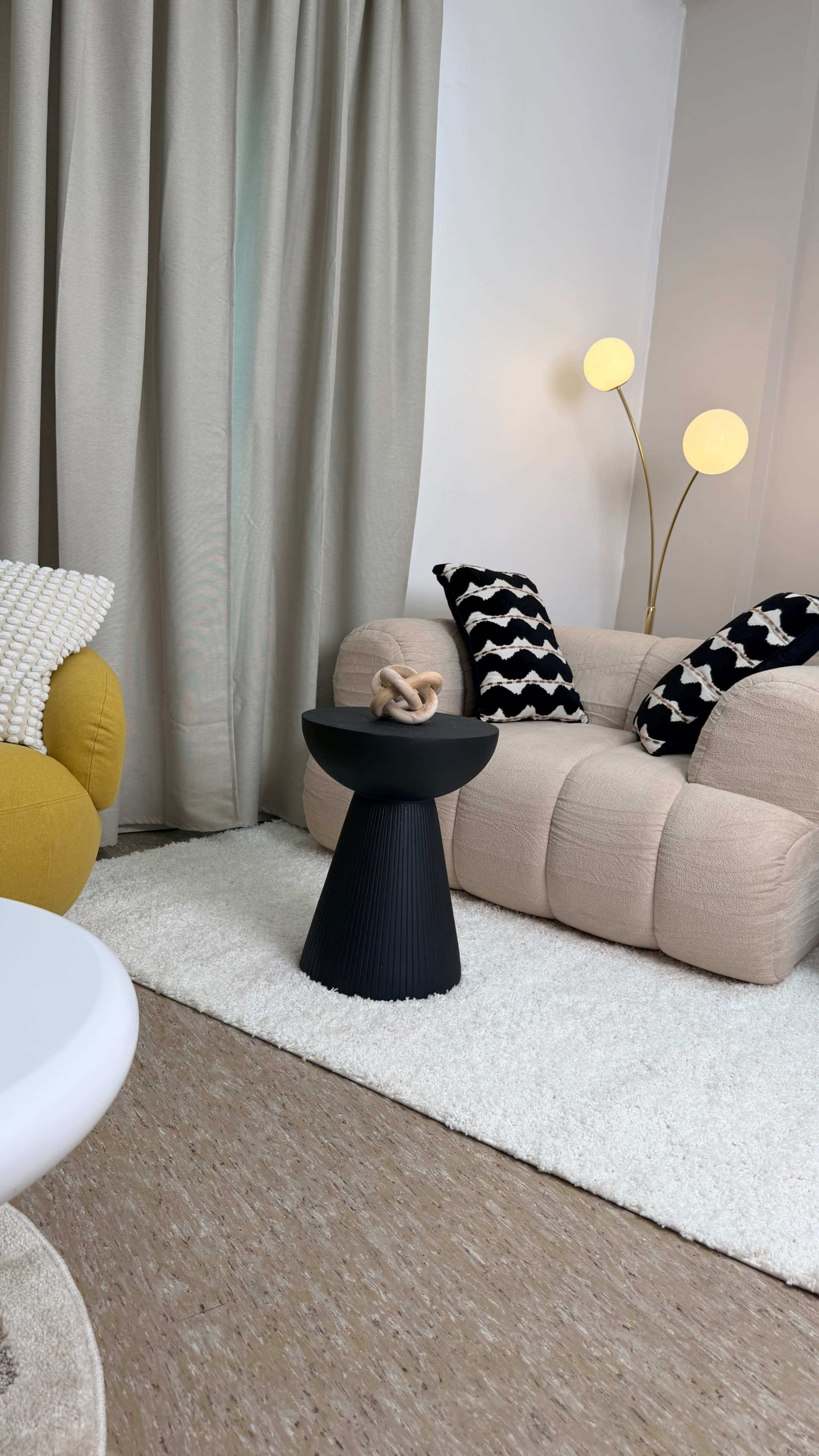 The scene features a cozy living room with a yellow armchair, a beige sofa, a black side table holding a decorative item, and soft, neutral-toned rugs and curtains.