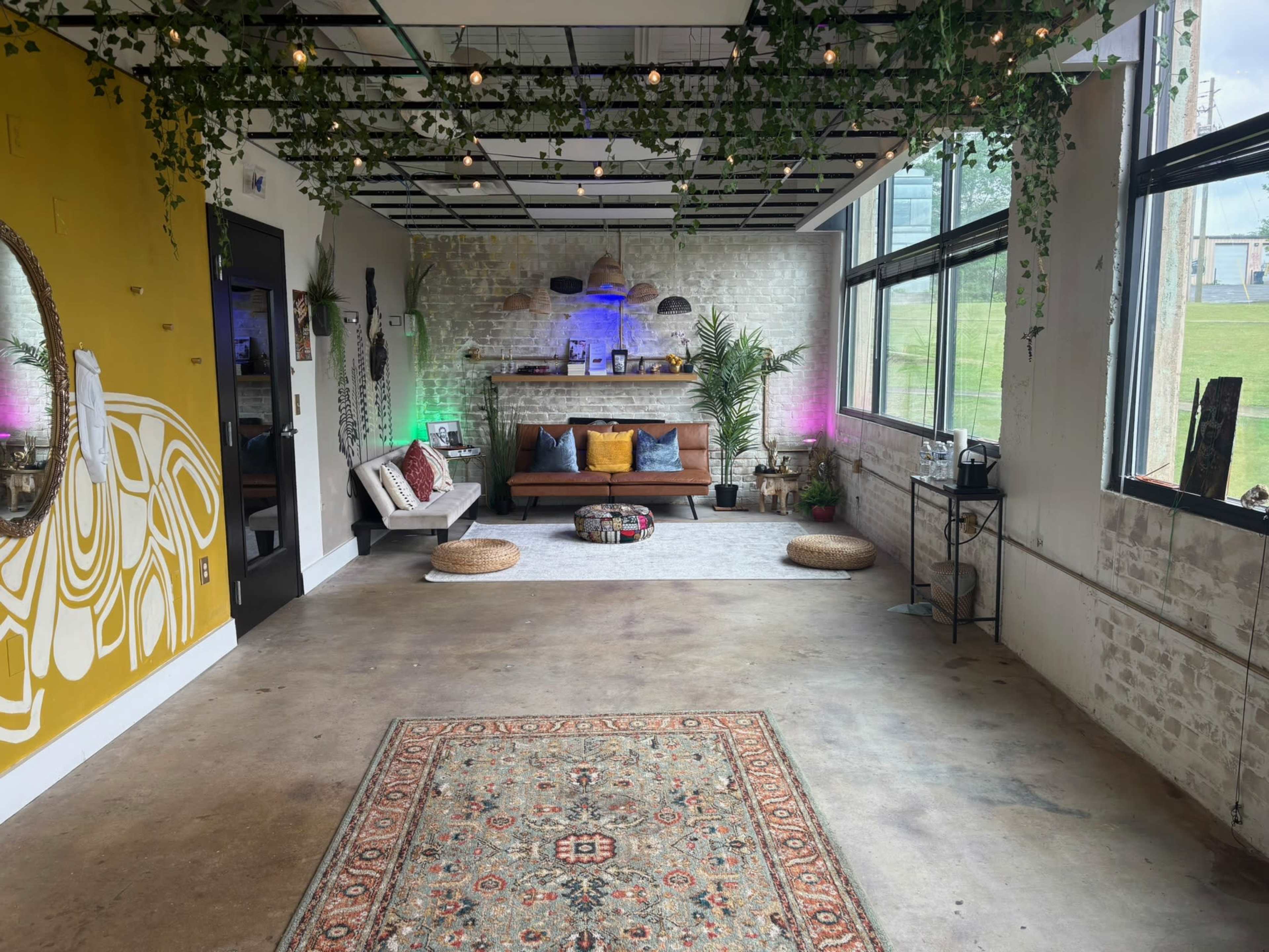The image shows a spacious, modern lounge area with a decorative rug, a couch against a textured wall, plants, and colorful lighting.