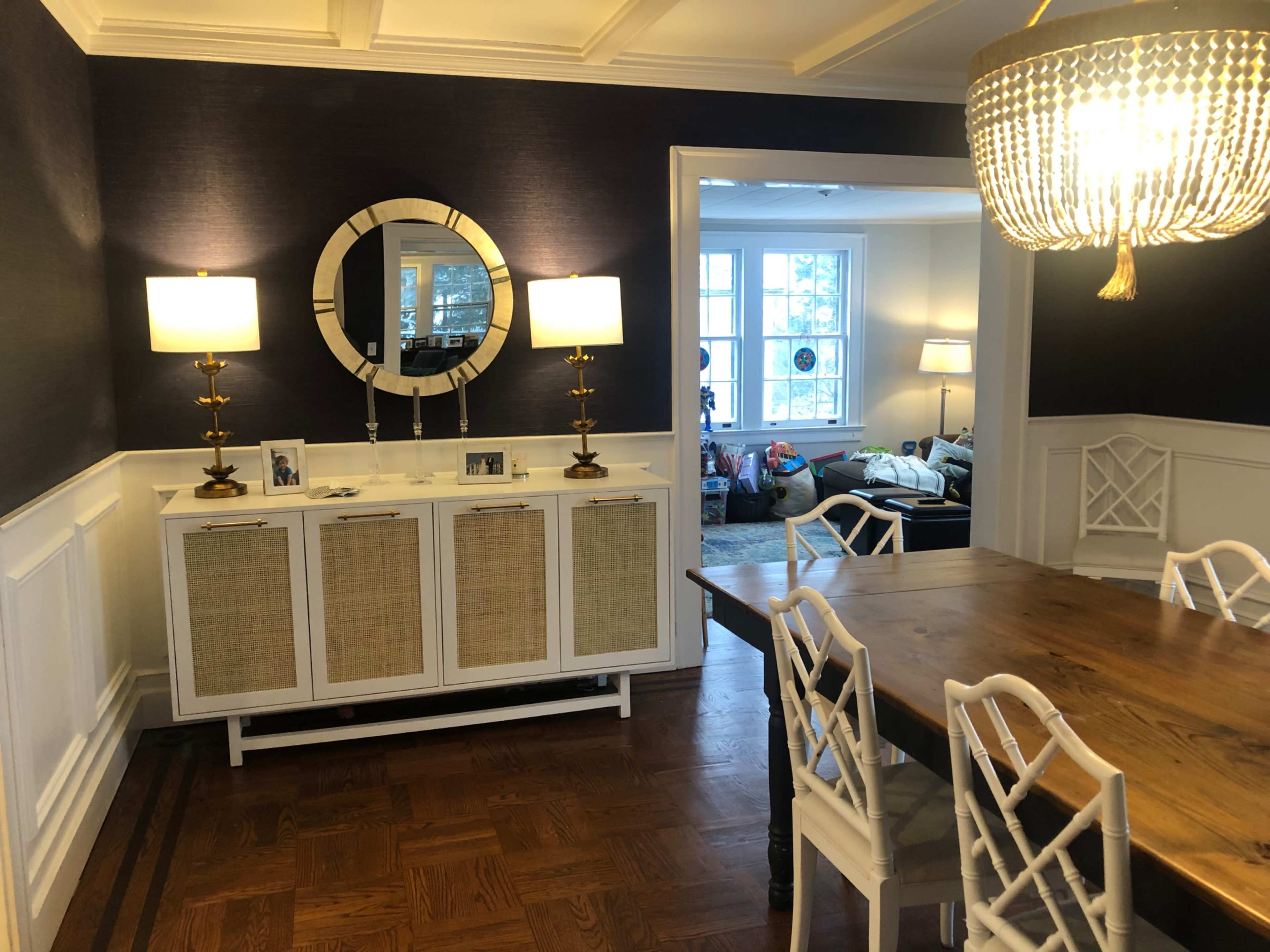 The image shows a dining area with a wooden table and white chairs, a sideboard with photographs, a round mirror on the wall, and a large chandelier illuminating the space.