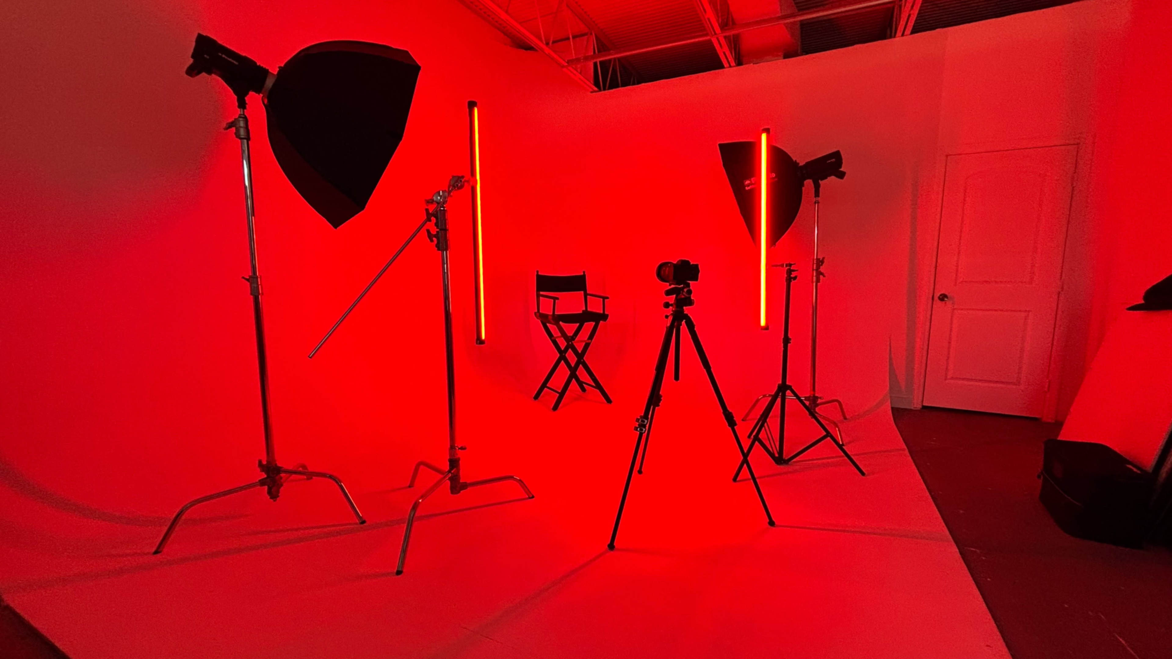The image shows a photography studio illuminated with red lighting, featuring various light stands, a camera on a tripod, and a director's chair.