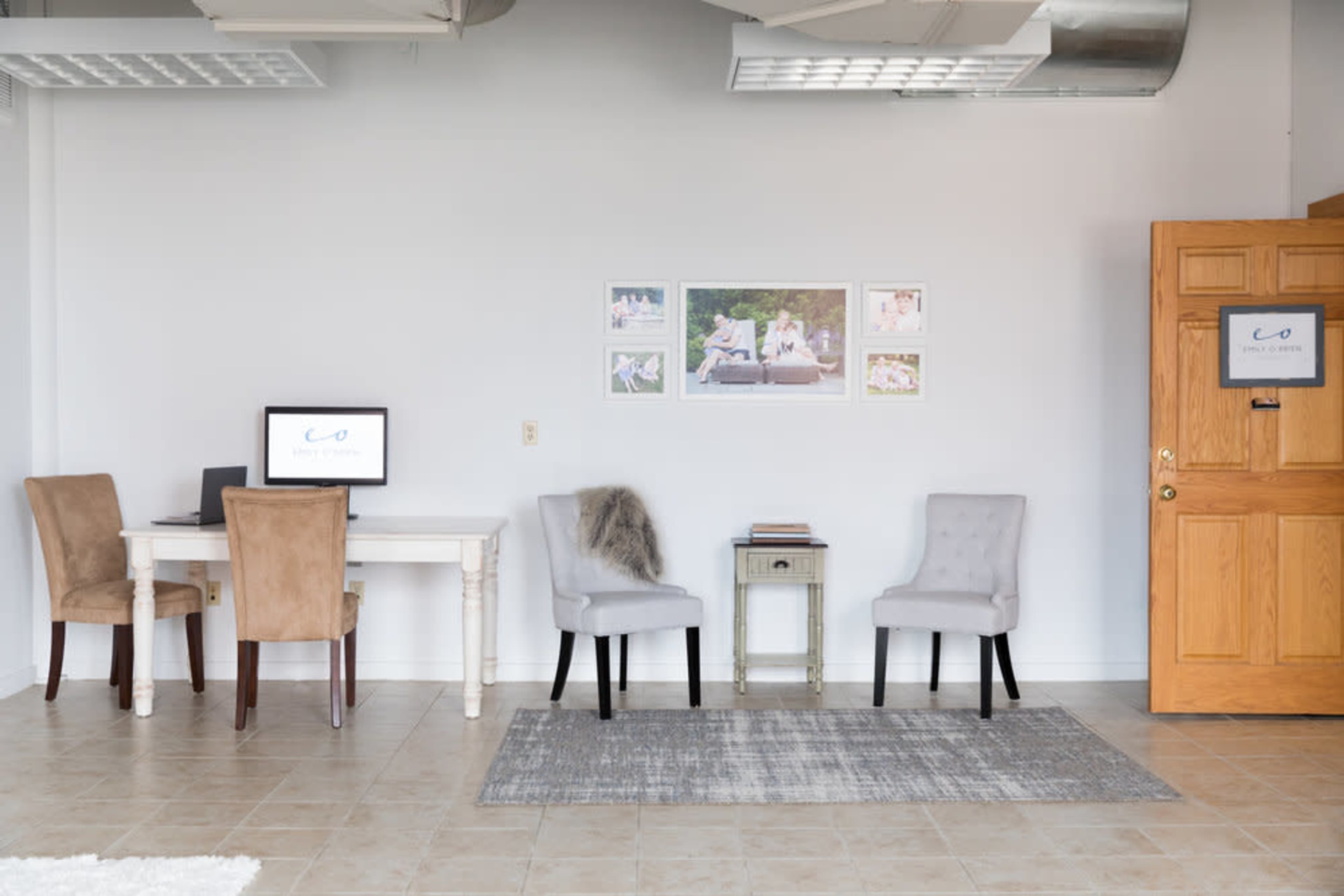 A modern office space with a desk, computer, and various chairs arranged alongside a door and framed photos on the wall.