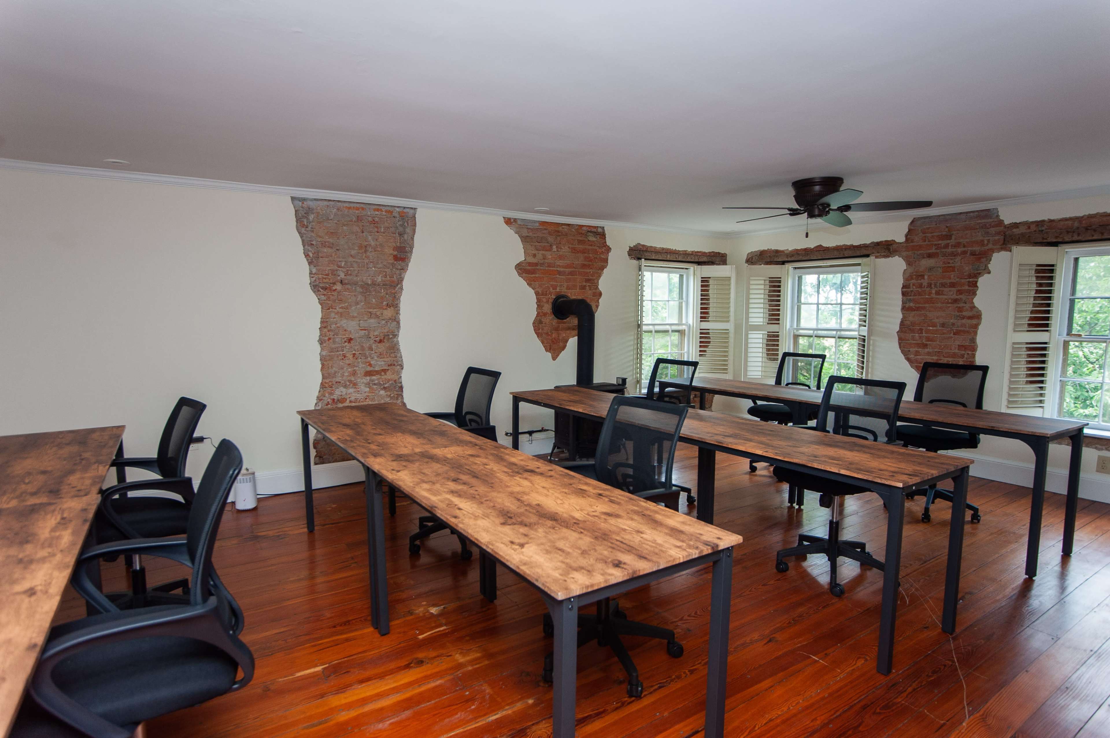 A spacious room with multiple wooden tables and black chairs arranged neatly, featuring exposed brick walls and large windows.