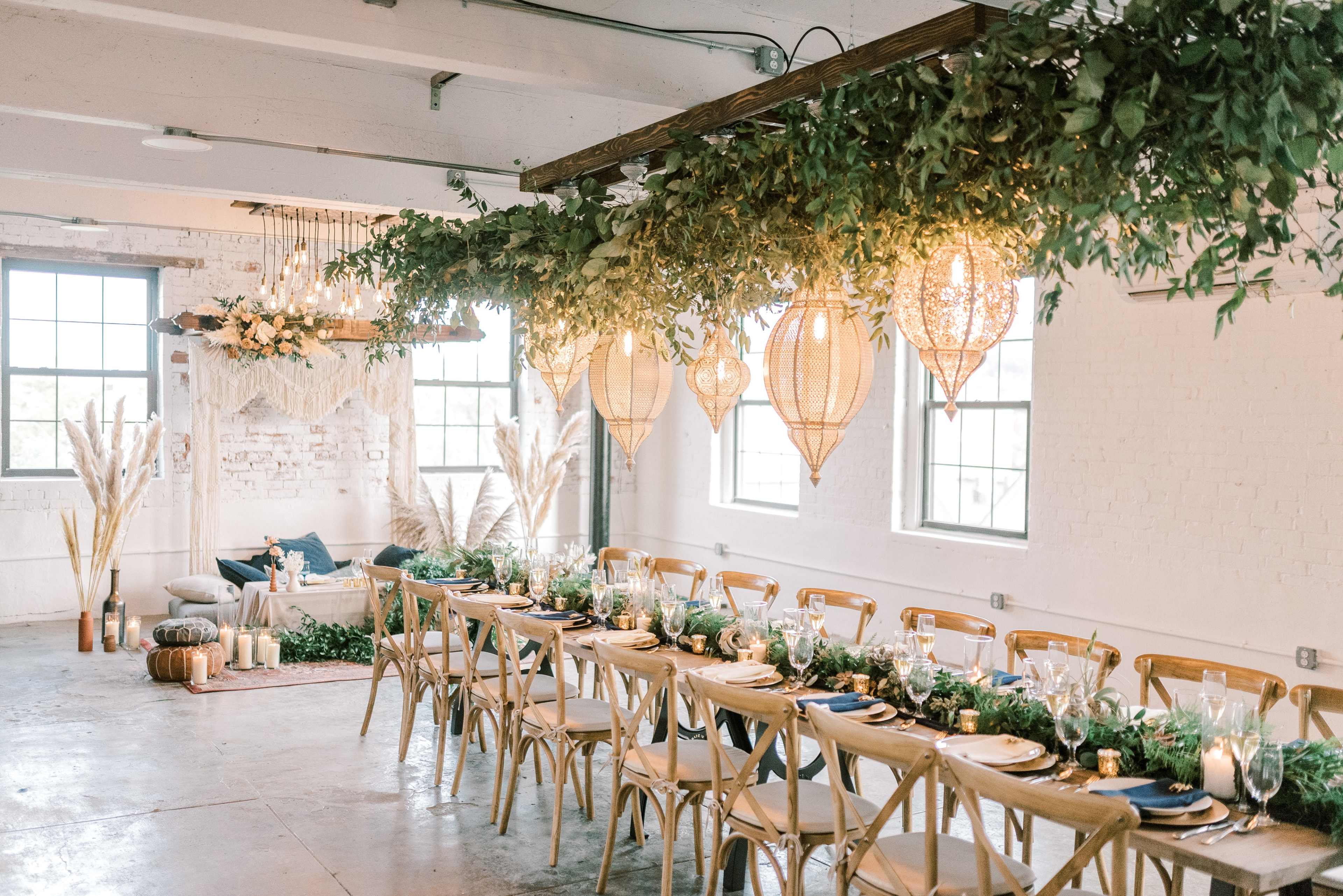A decorated dining area featuring a long table with elegant table settings, surrounded by greenery and hanging lanterns, in a bright, industrial-style room with large windows.