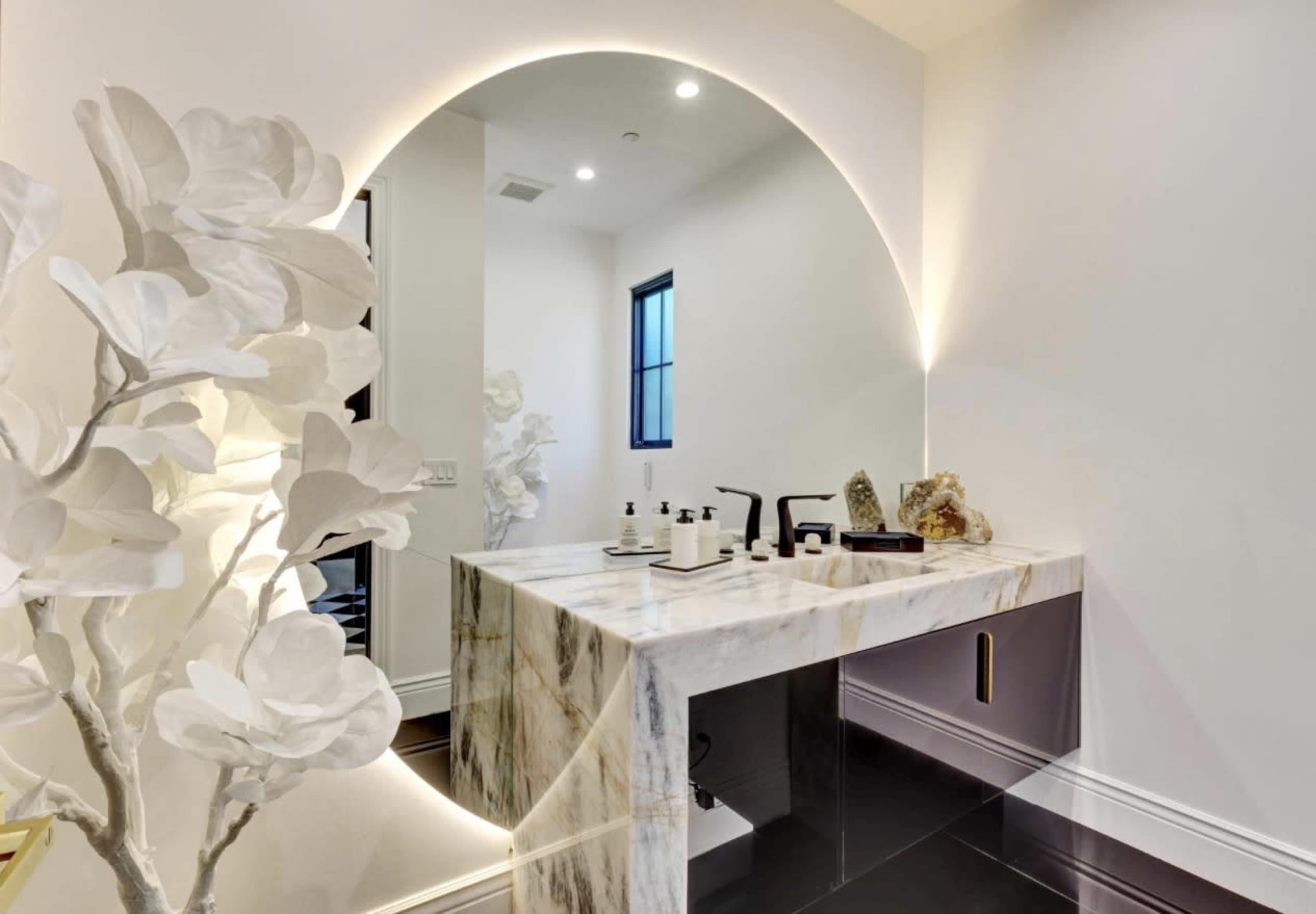 The image depicts a modern bathroom with a marble vanity illuminated by a sleek, curved mirror and accompanied by decorative white plants.