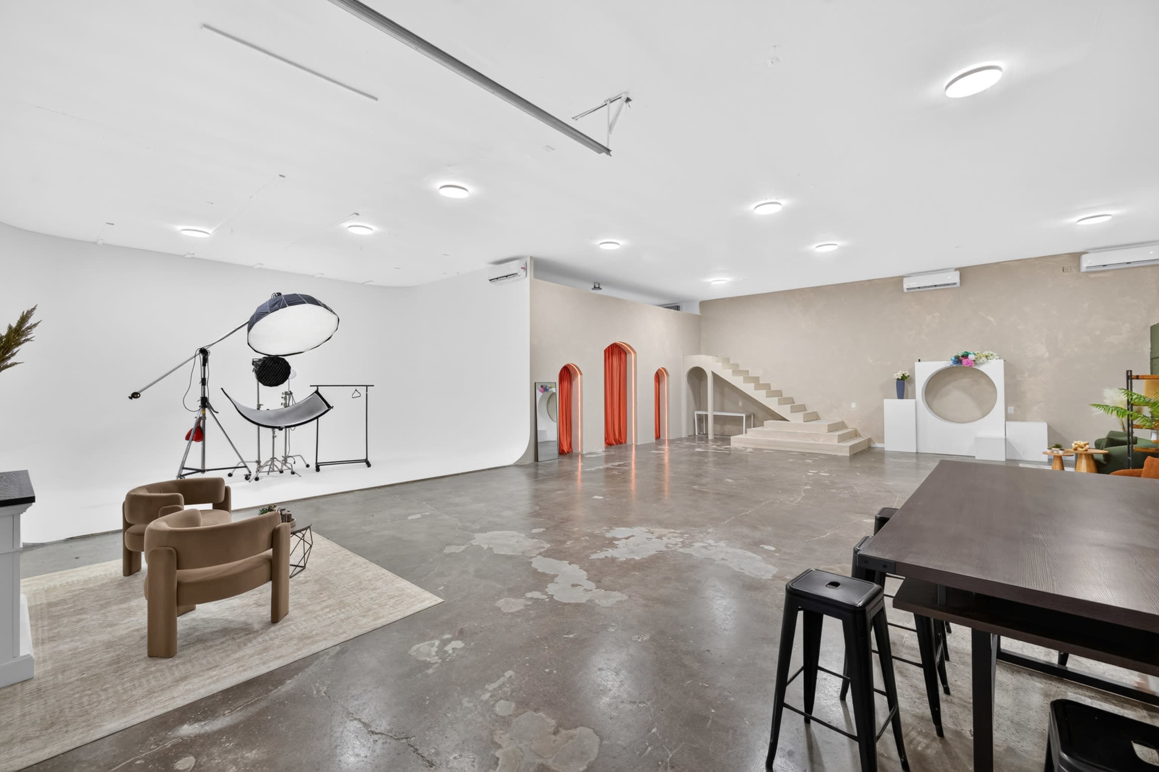 The image shows a spacious studio interior featuring a white backdrop, beige walls, modern furniture, and stairs leading to an upper level.