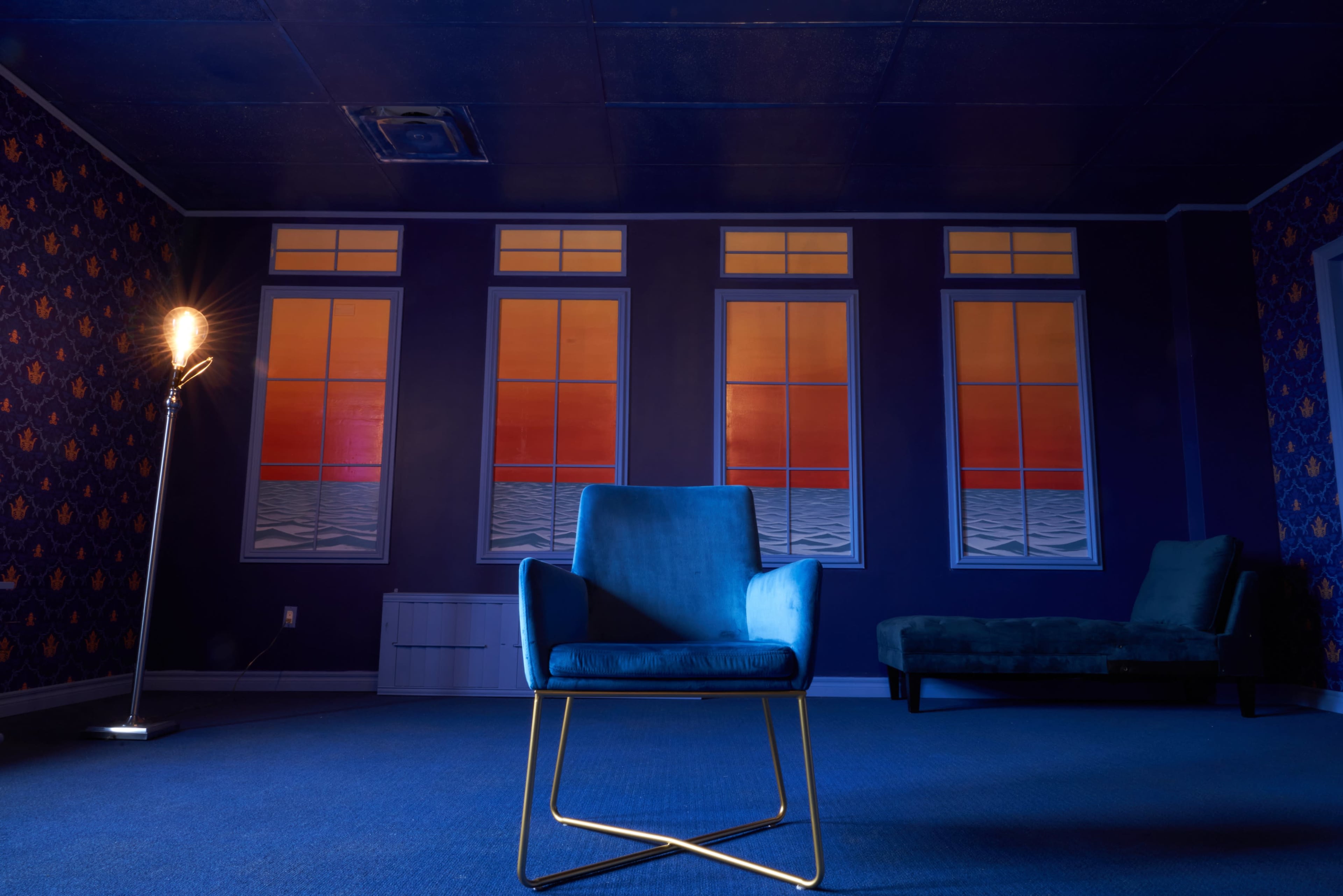 A blue chair sits in a dimly lit room with patterned walls and large windows displaying a colorful sunset.