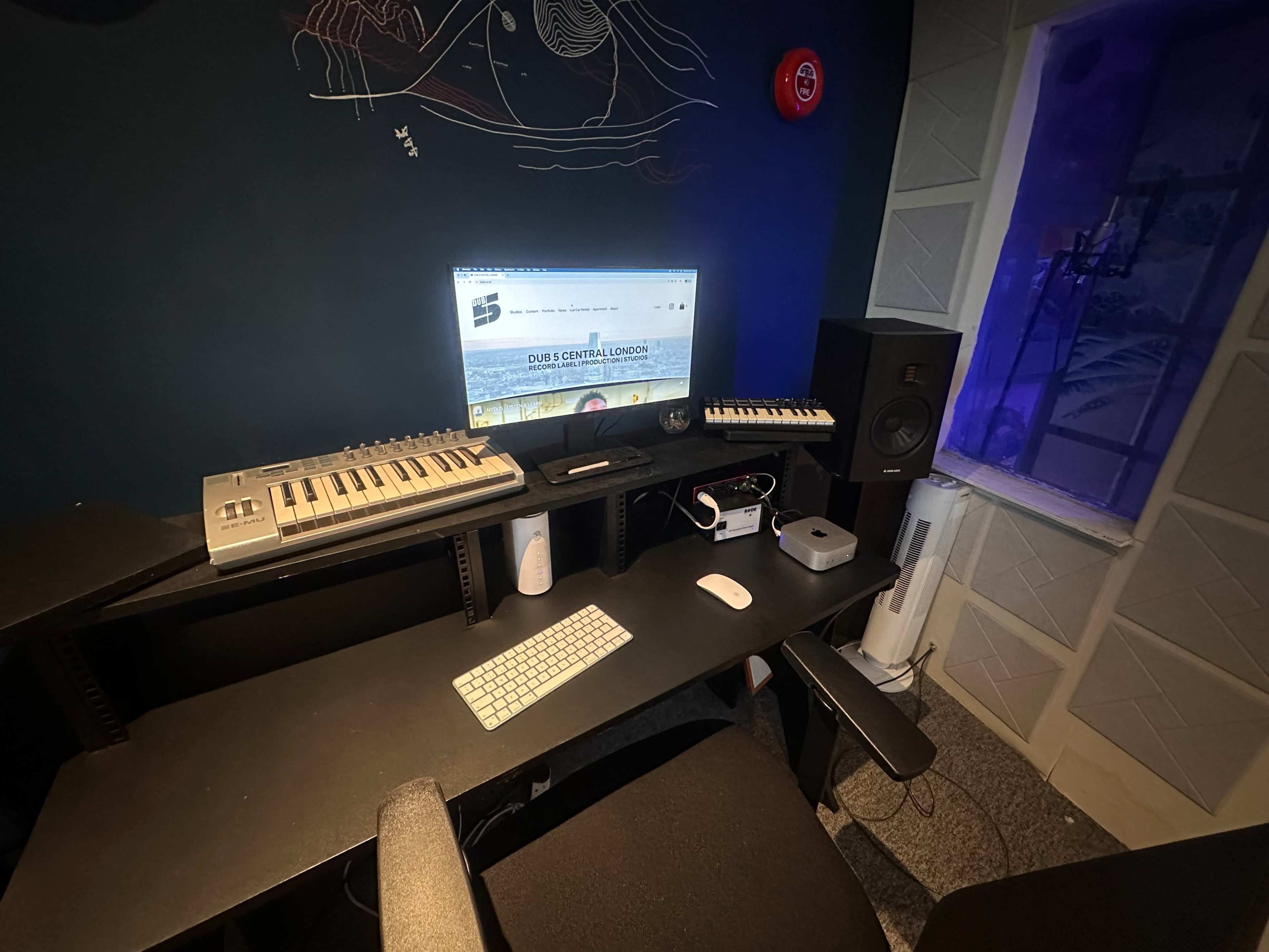The image shows a music production workspace with a computer, keyboard, audio monitors, and a window overlooking a purple-lit area.