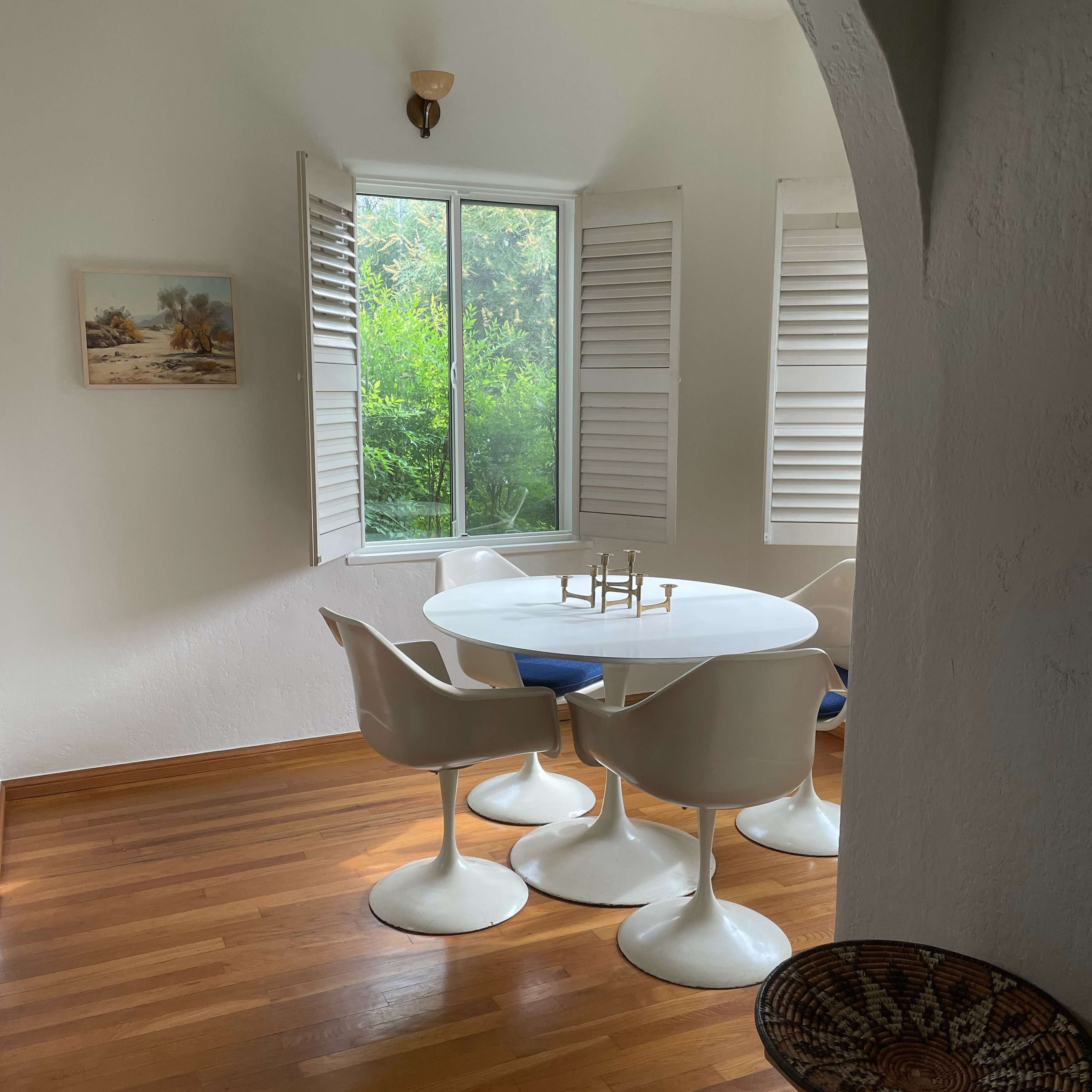 A dining area features a round white table with four white chairs, positioned near a window with open shutters, allowing natural light to illuminate the wooden floor.