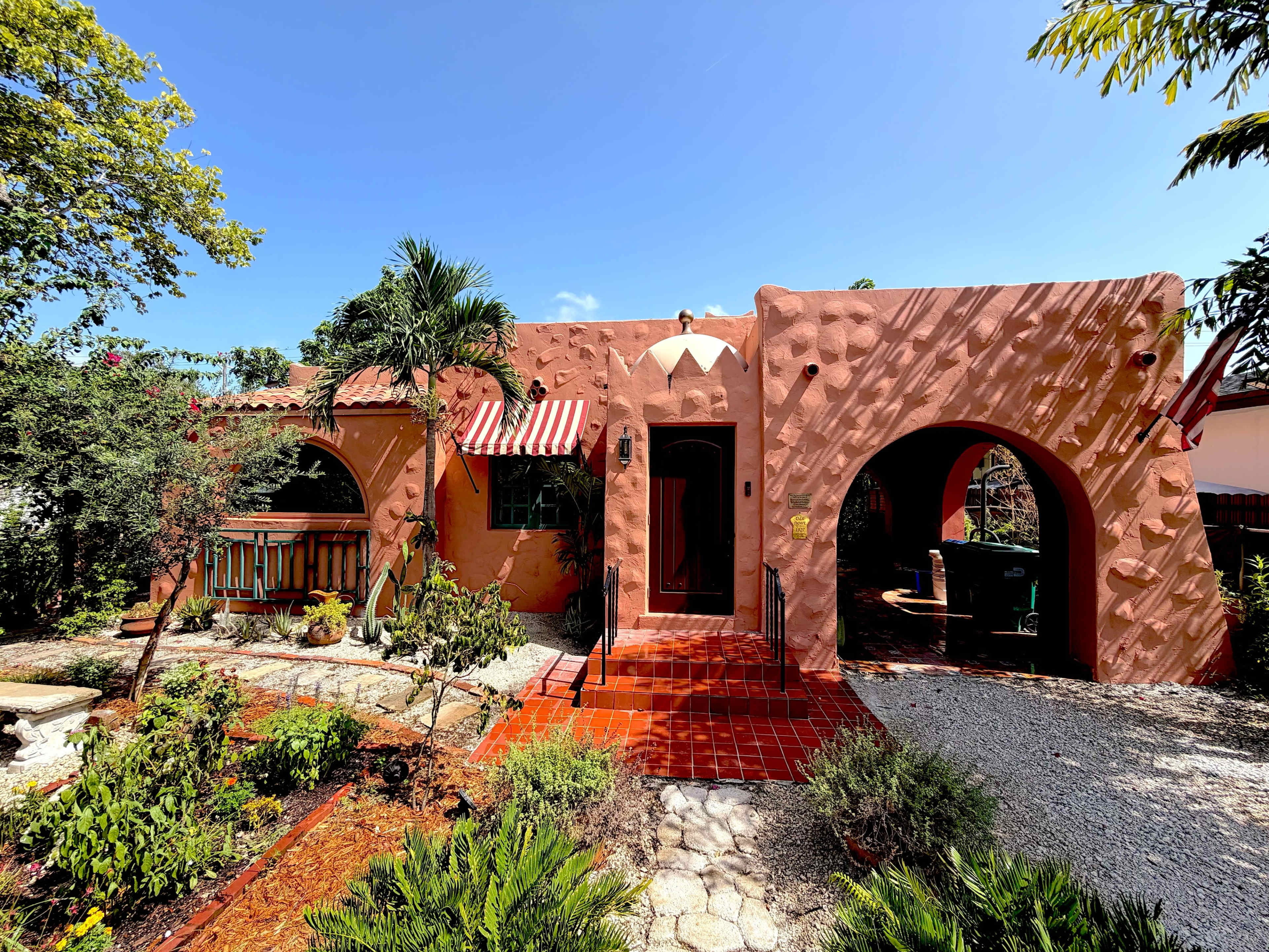 A colorful adobe-style house features multiple archways, red tile accents, and landscaped gardens.