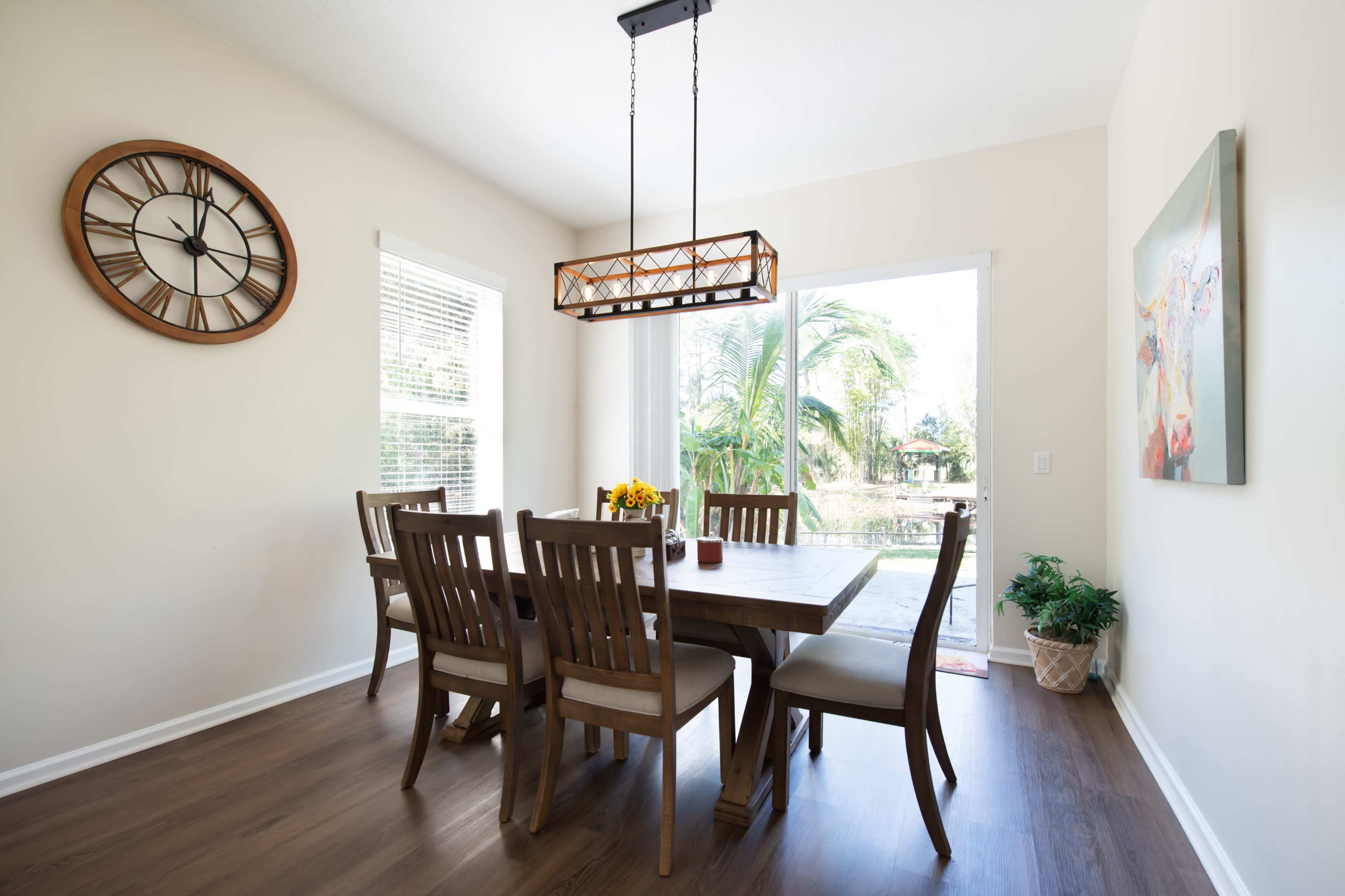 A spacious dining area features a wooden table surrounded by six chairs, a large clock on the wall, and a sliding door leading to a patio with greenery outside.