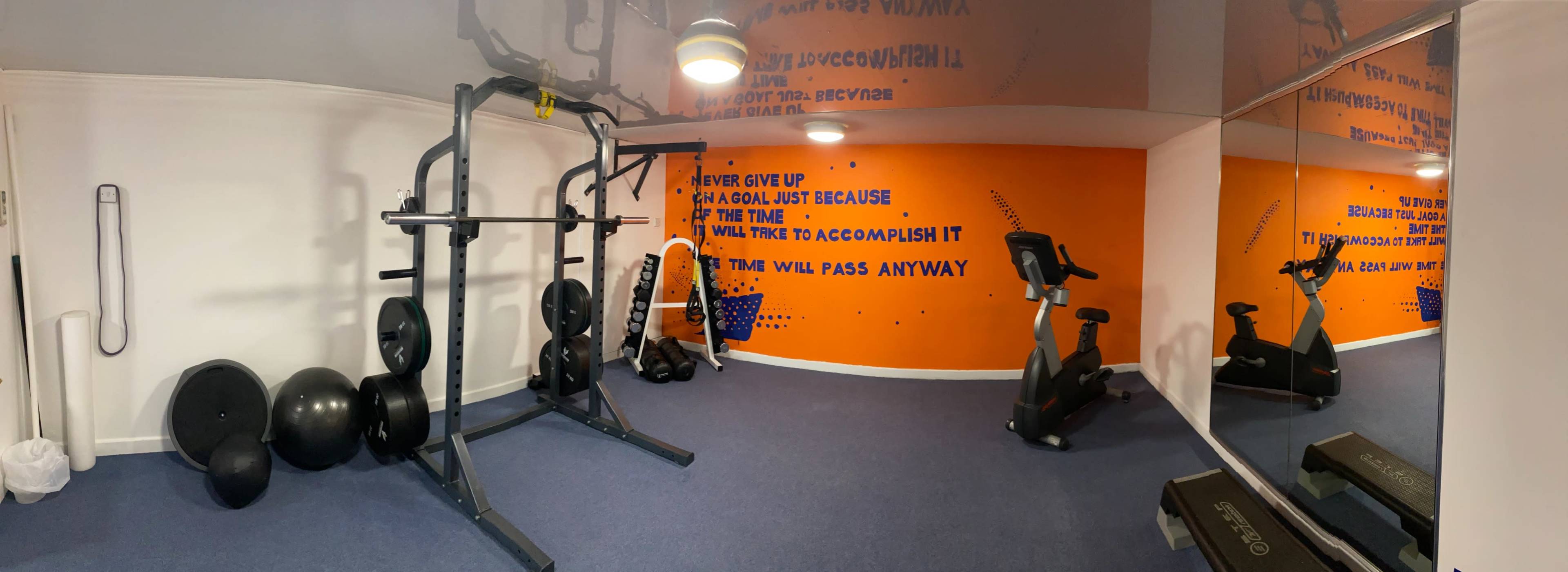The image shows a compact gym space featuring exercise equipment such as a weight rack, exercise bikes, stability balls, and motivational wall art.