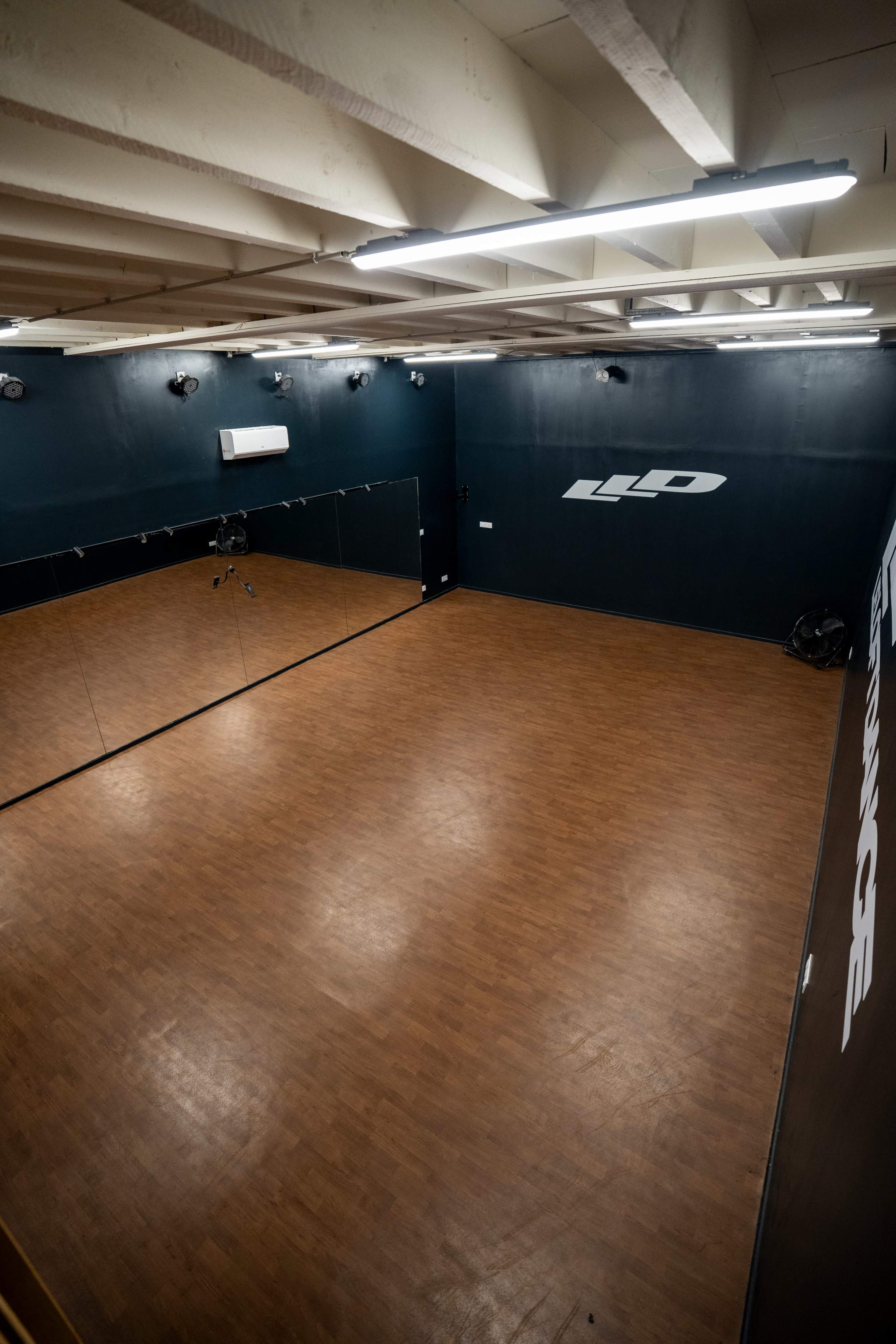 The image shows a spacious dance or rehearsal studio with wooden flooring, mirrored walls, and minimal lighting fixtures.