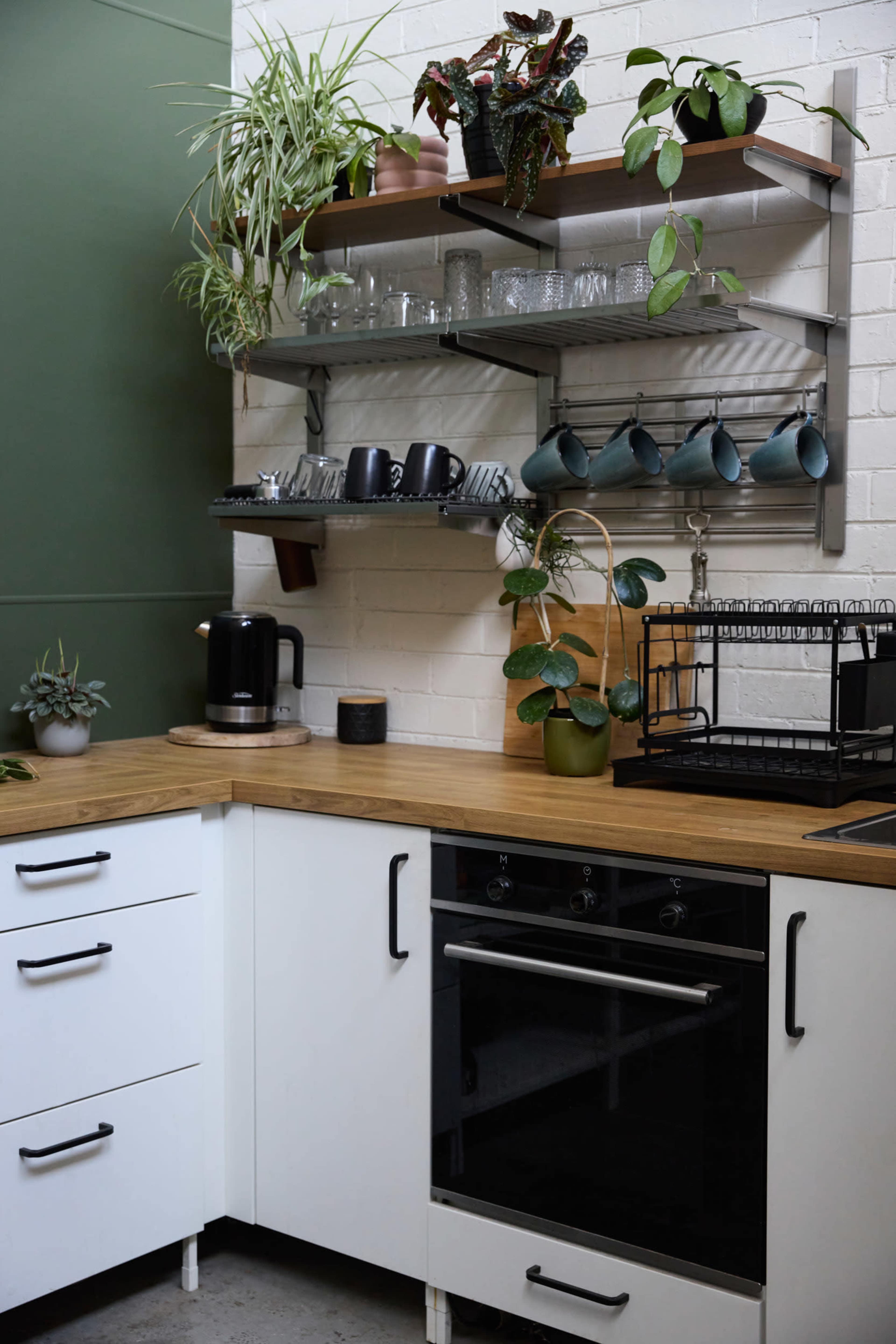 A modern kitchen features a wooden countertop, black oven, and open shelves with plants and glassware against a green accent wall.