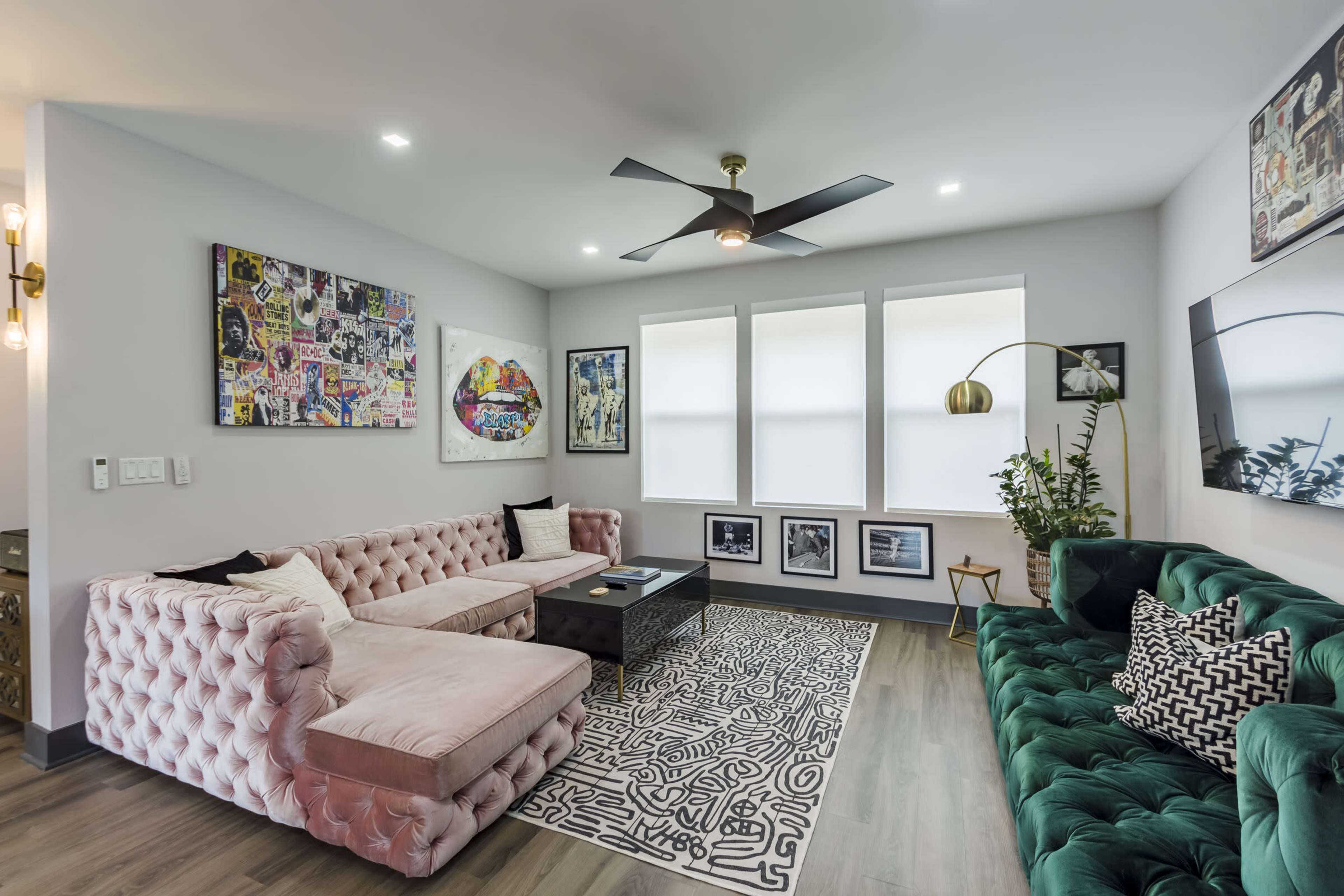 The image shows a modern living room featuring a pink tufted sectional sofa, a green velvet sofa, a black coffee table, and large windows adorned with white blinds, along with colorful wall art.