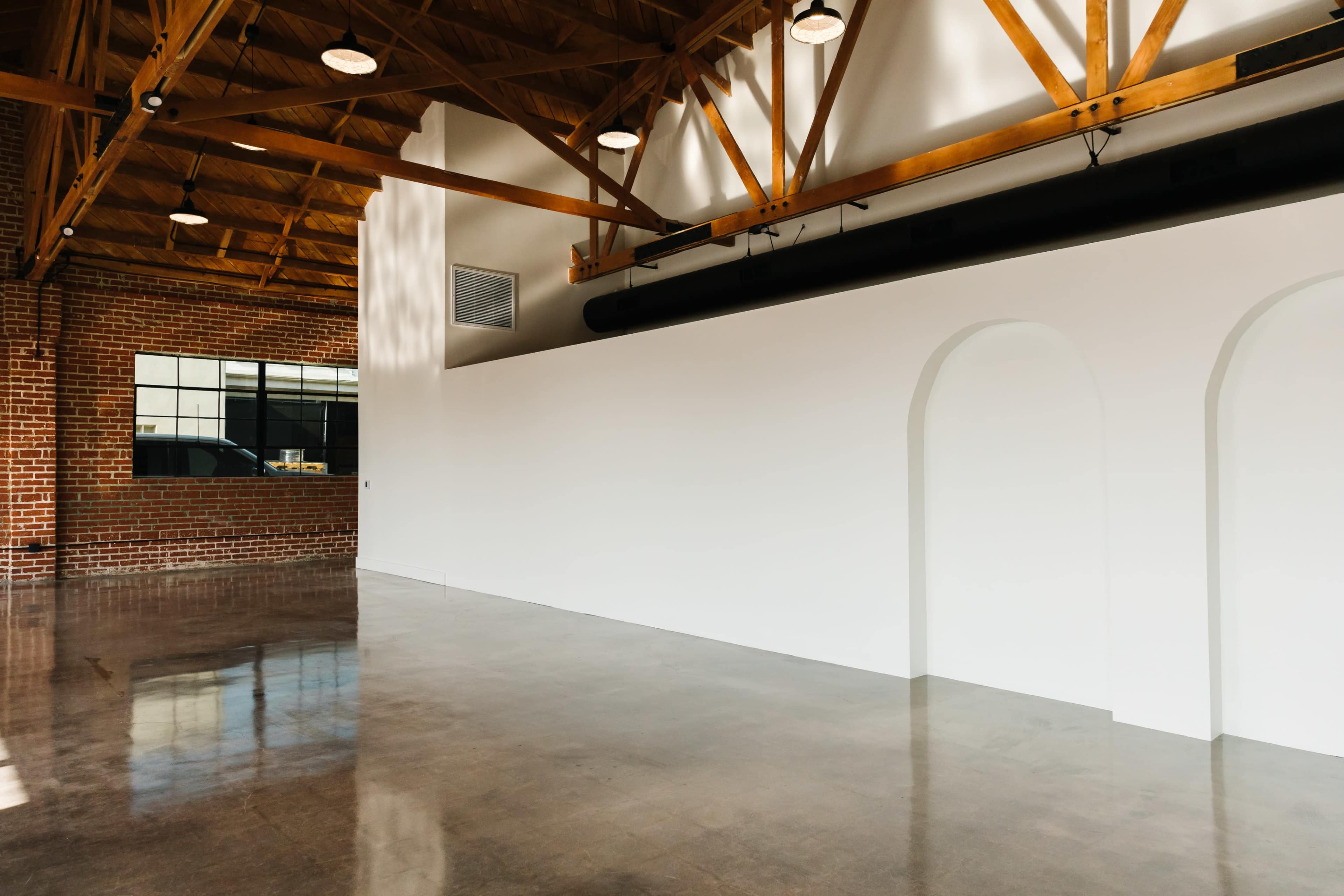 A spacious interior with a polished concrete floor, exposed wooden beams, and an empty white wall featuring an arched opening.