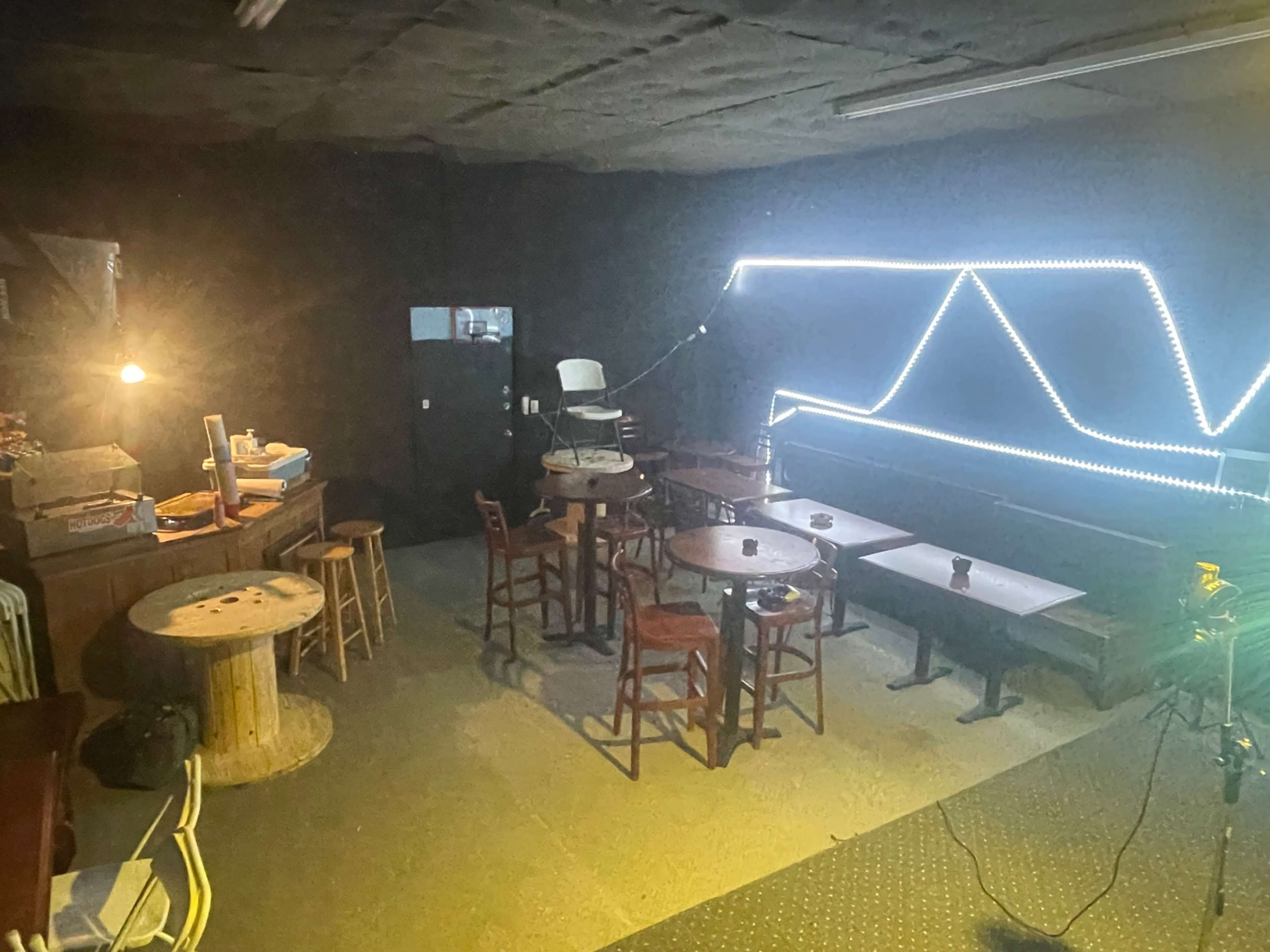 The image shows a dimly lit room with several wooden tables and chairs, a bar area in the corner, and decorative light strings along the wall.