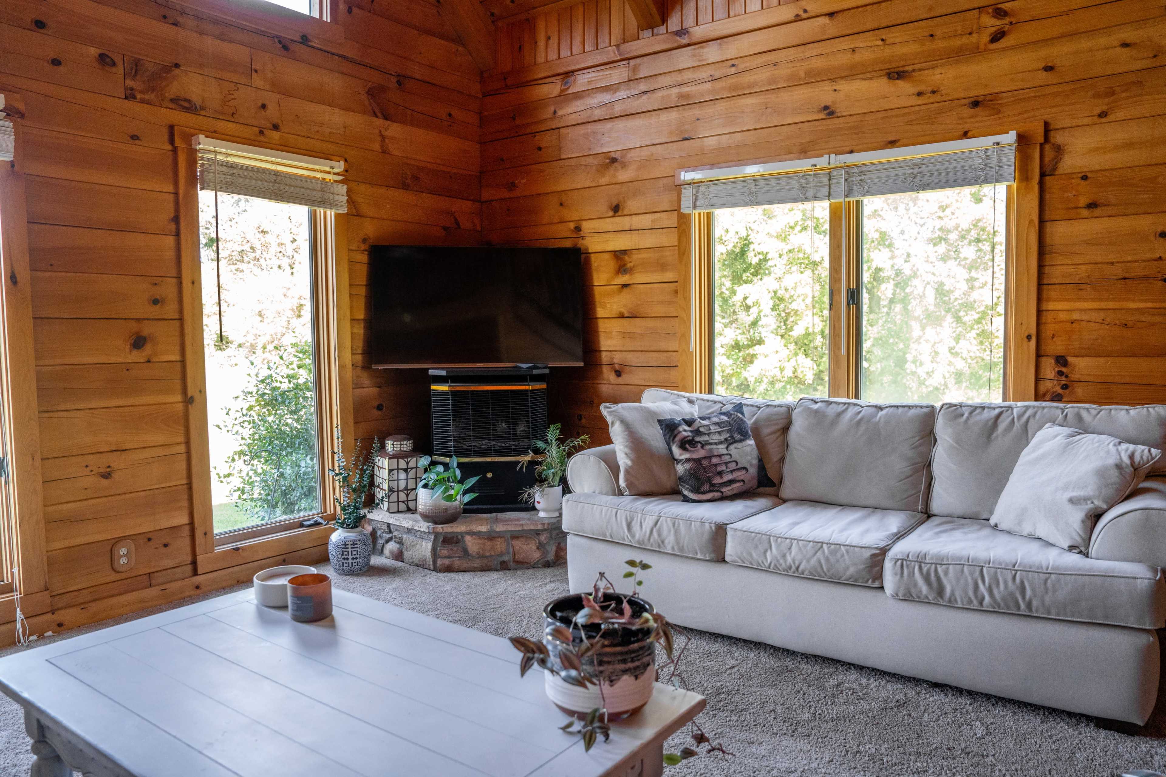 A cozy living room with wooden walls, a beige sofa, a coffee table, and a television mounted on the wall beside large windows.
