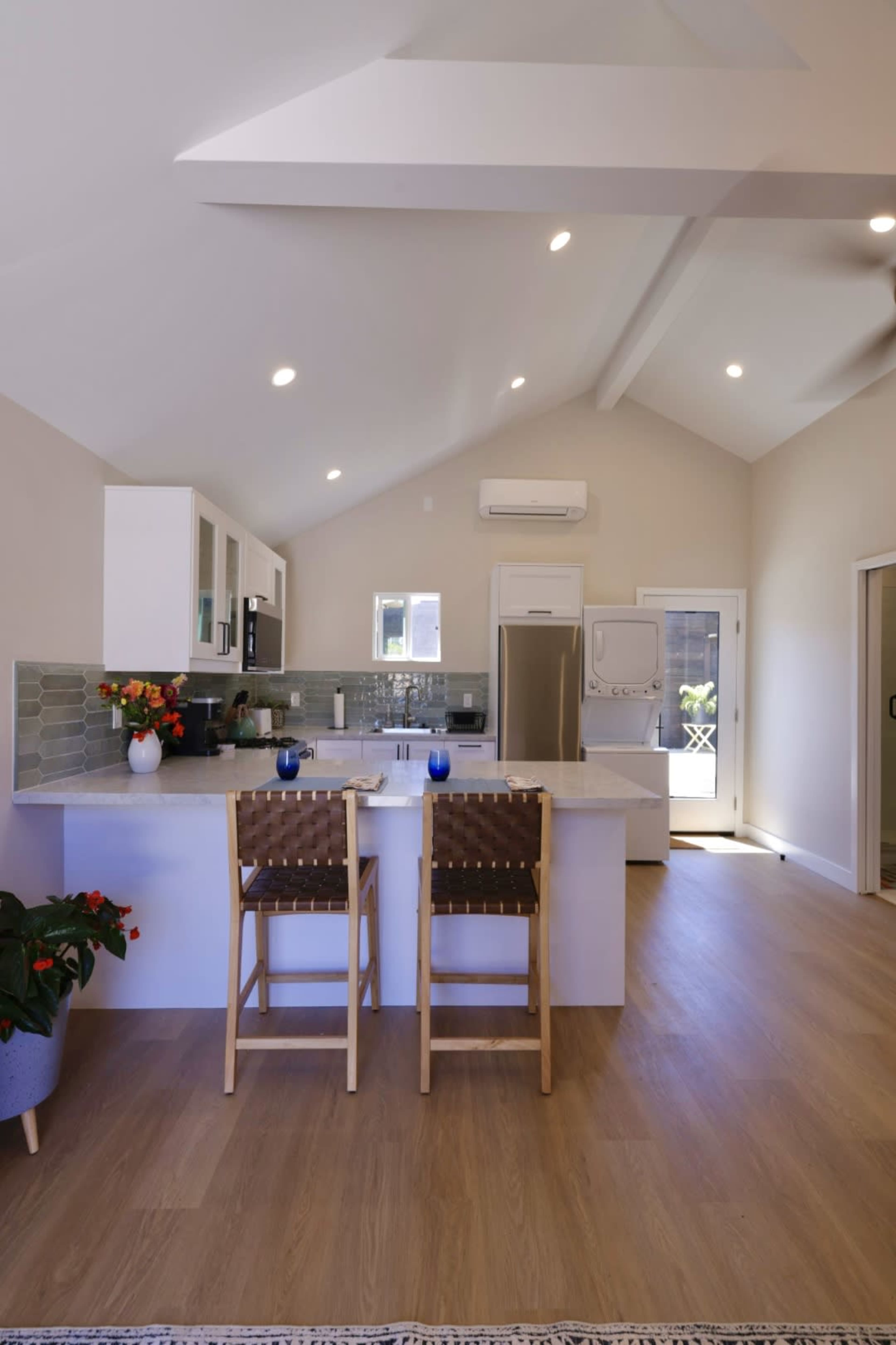 A bright kitchen features a white island with two wooden barstools, modern appliances, and large windows allowing natural light.