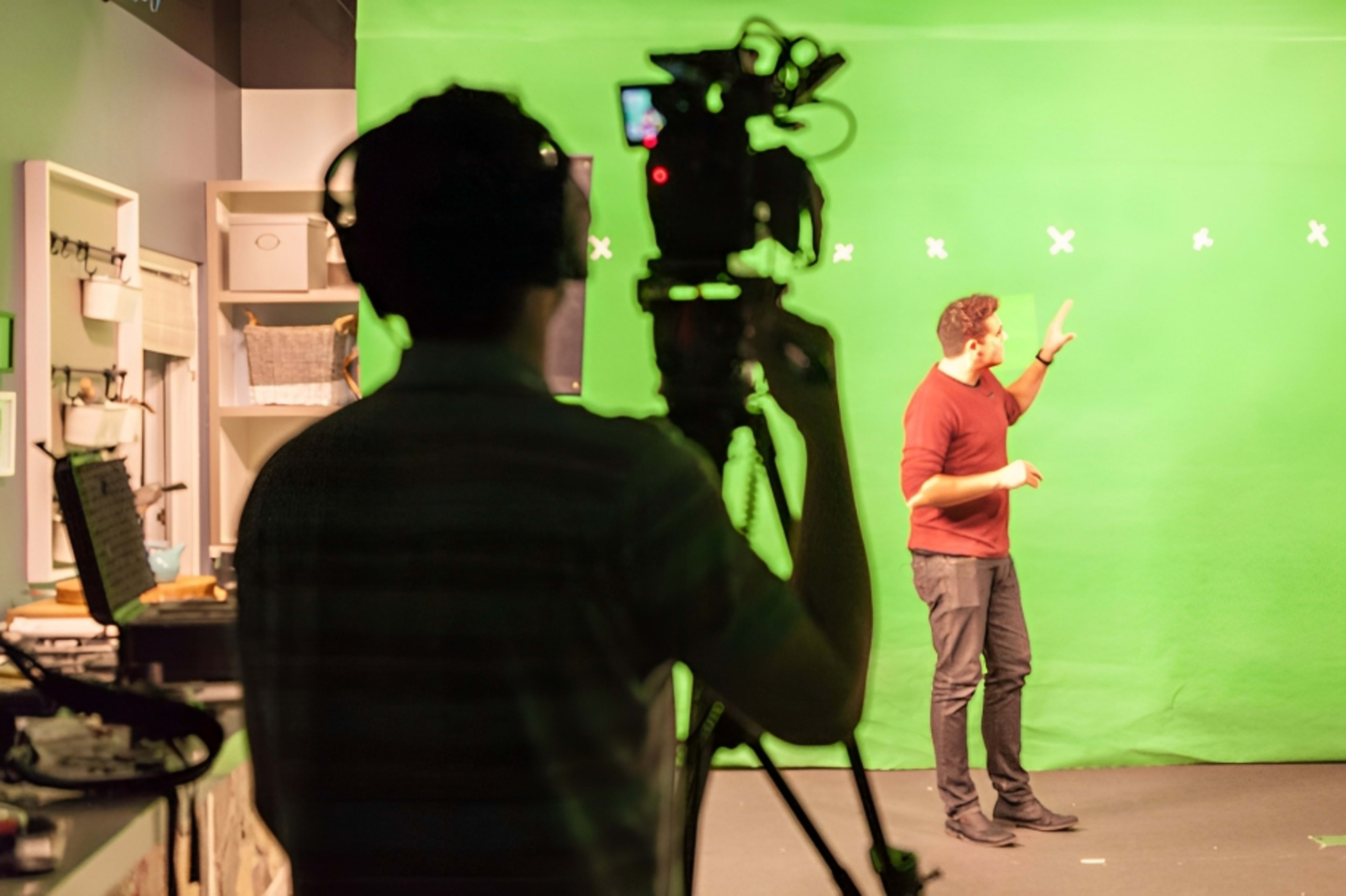 A person gestures in front of a green screen while another individual operates a camera from the foreground.