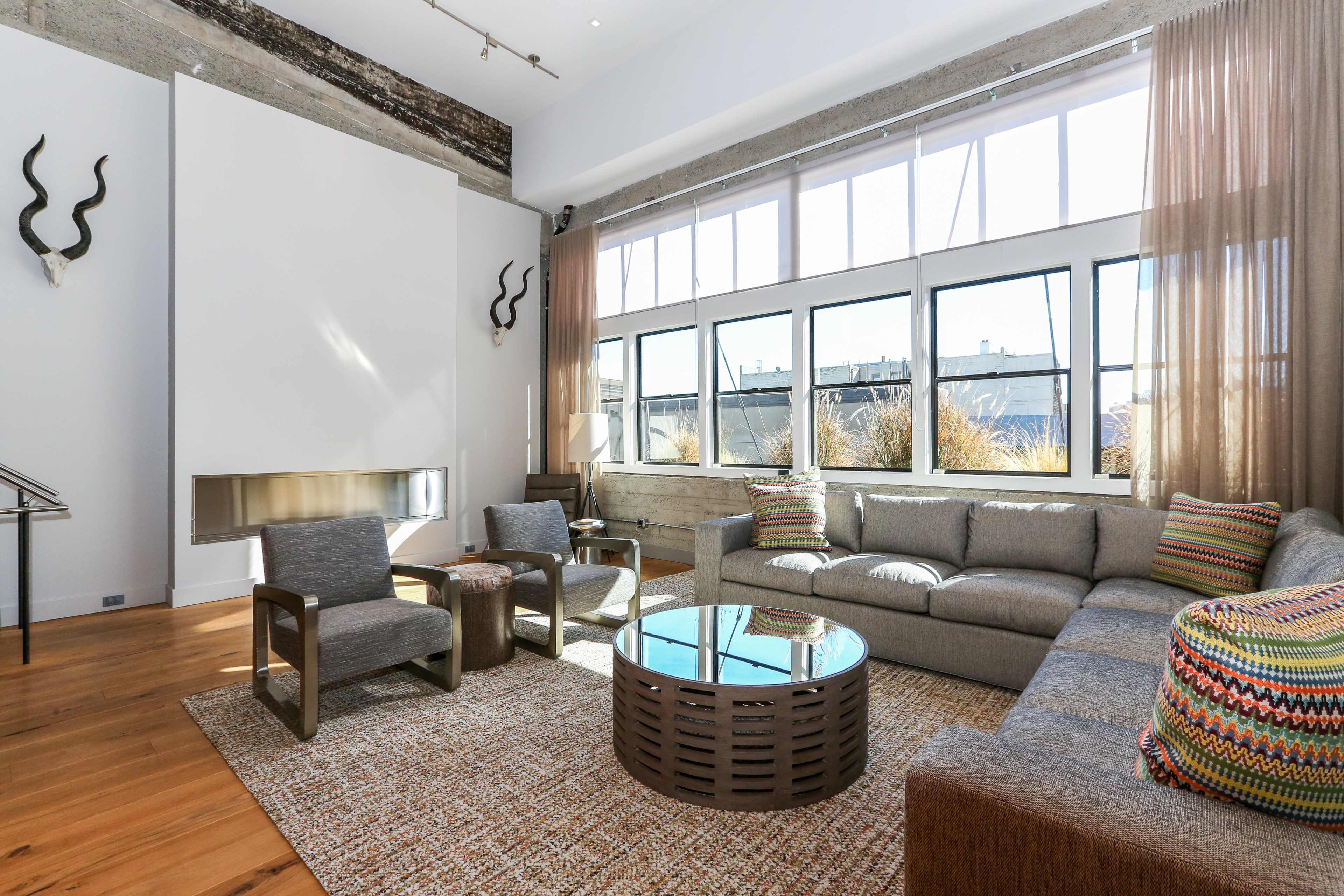 A modern living room with large windows, a gray sofa, a round coffee table, and decorative elements like animal horns on the walls.