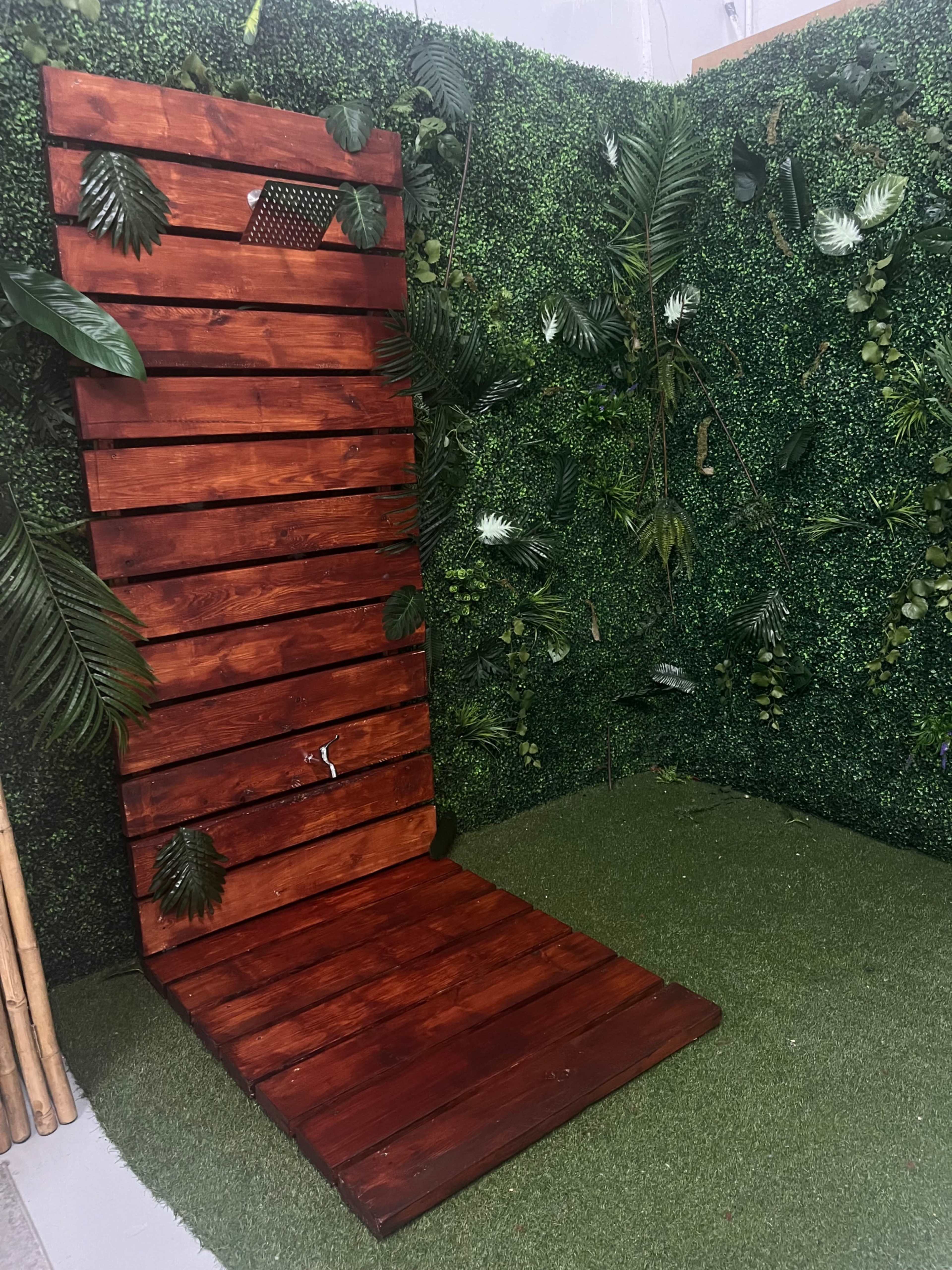 The image shows a wooden shower with slatted sides, featuring a shower head and surrounded by green foliage on a grass-like surface.