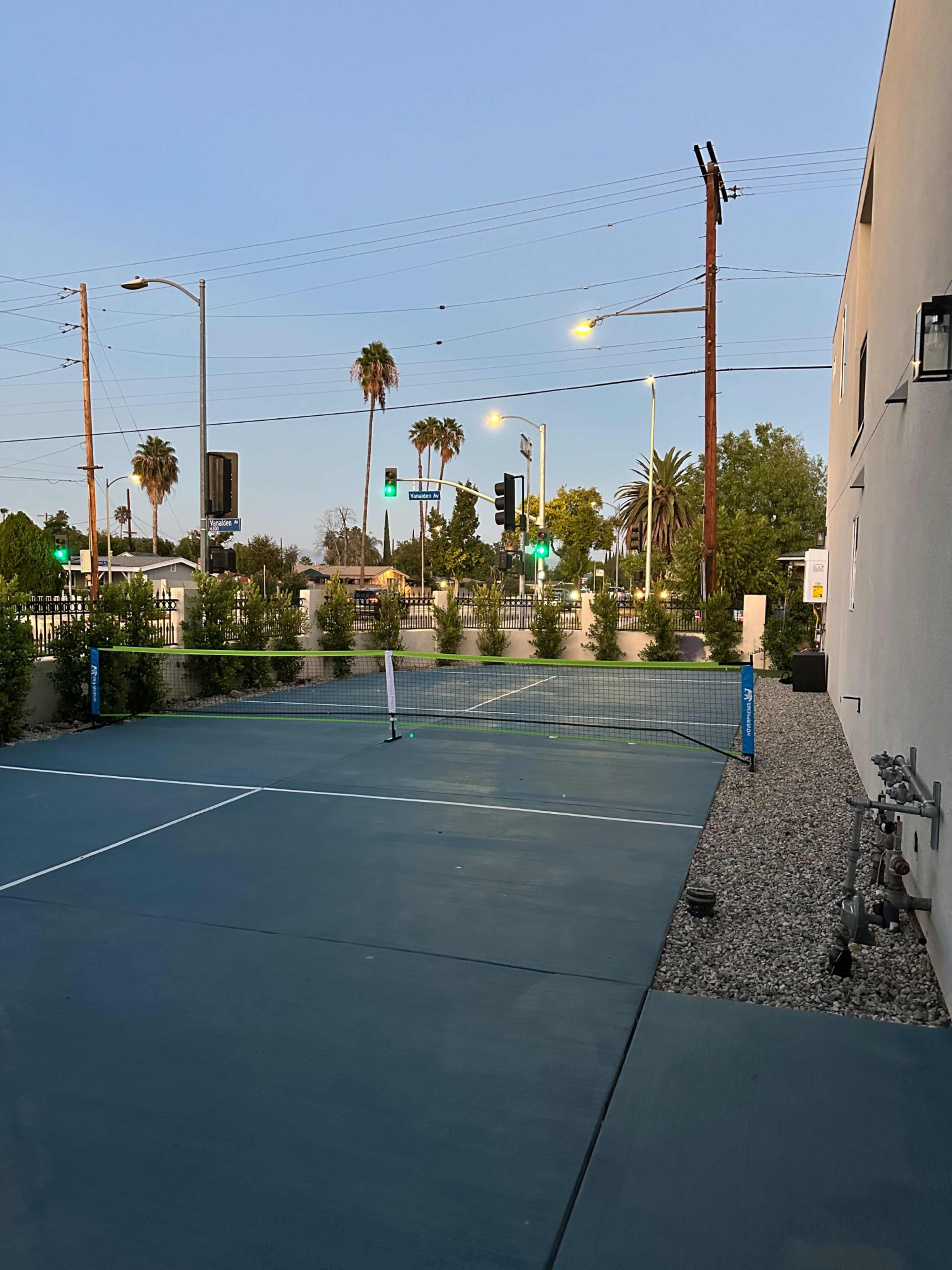 The image shows a tennis court with a net, surrounded by palm trees and streetlights, in an urban setting during dusk.