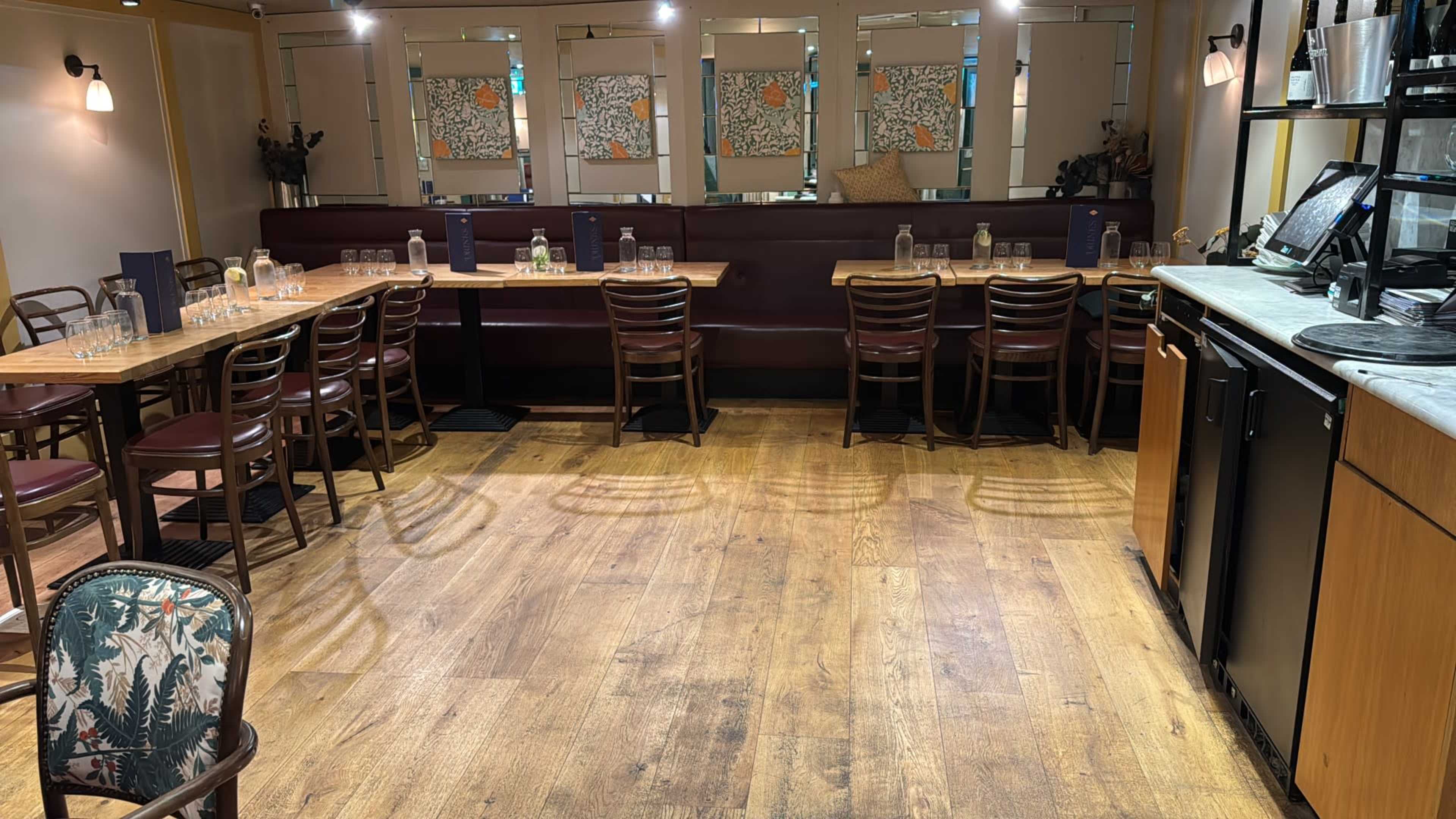 The image shows an empty restaurant interior with wooden floors, multiple tables and chairs arranged neatly, and decorative wall art in the background.