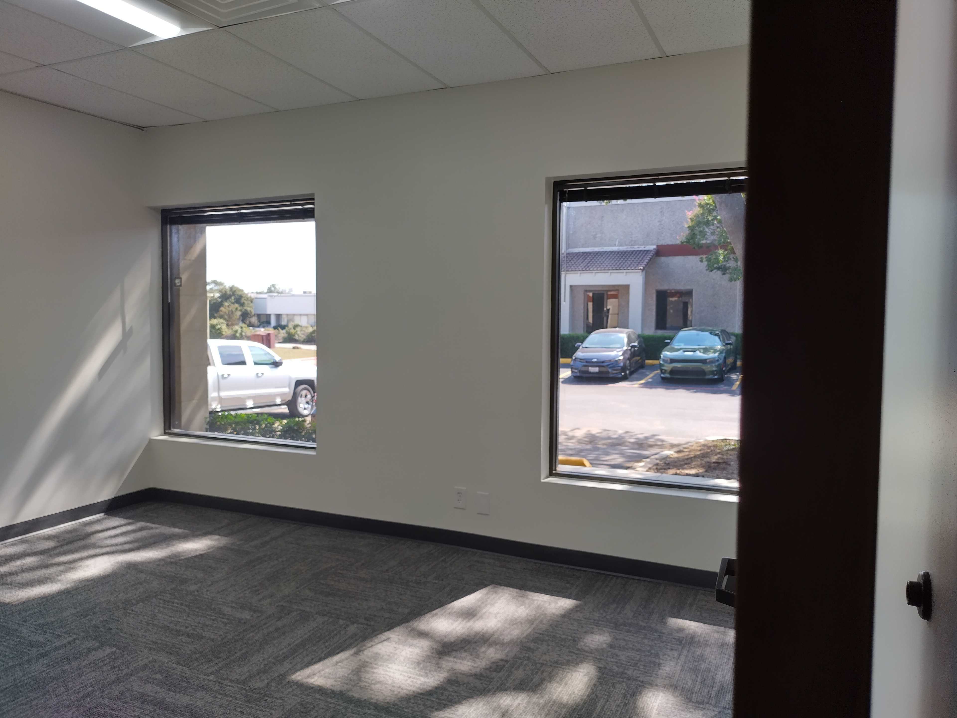 The image shows an empty office space with two large windows and a view of parked cars outside.