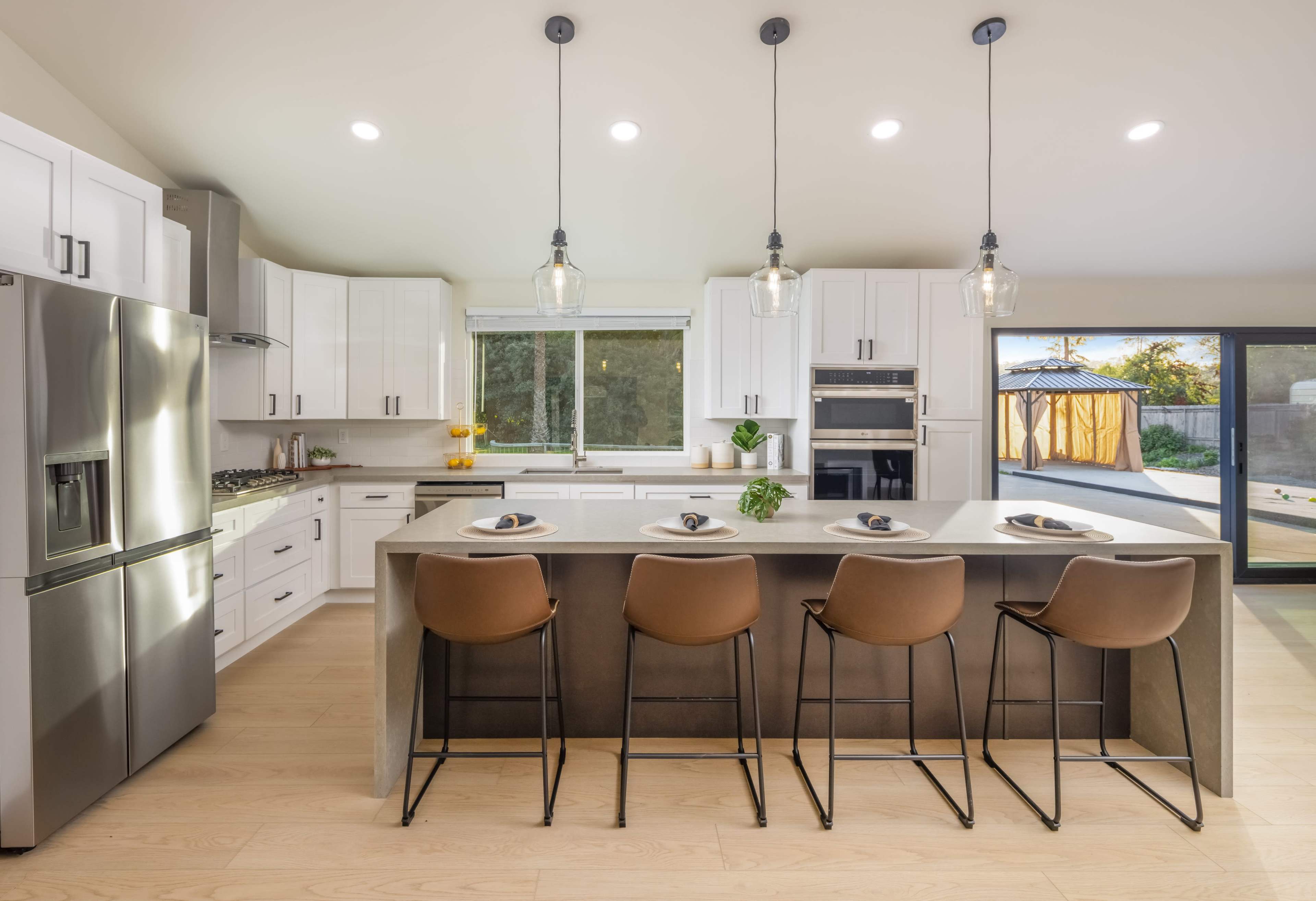 A modern kitchen features a concrete island with four brown bar stools, stainless steel appliances, and bright overhead lighting.