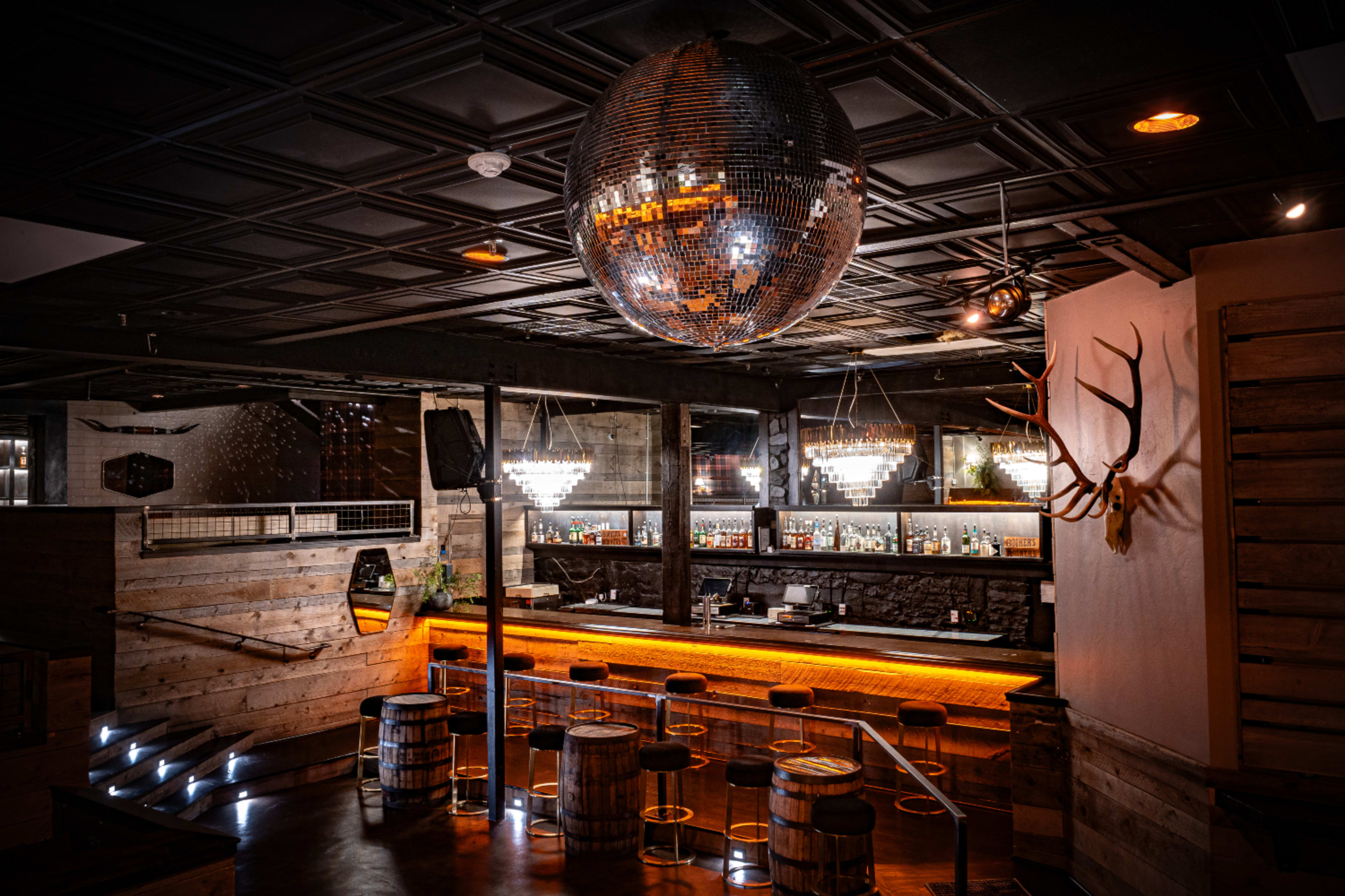 A dimly lit bar interior featuring a large disco ball overhead, wooden accents, and a well-stocked bar with stylish lighting.