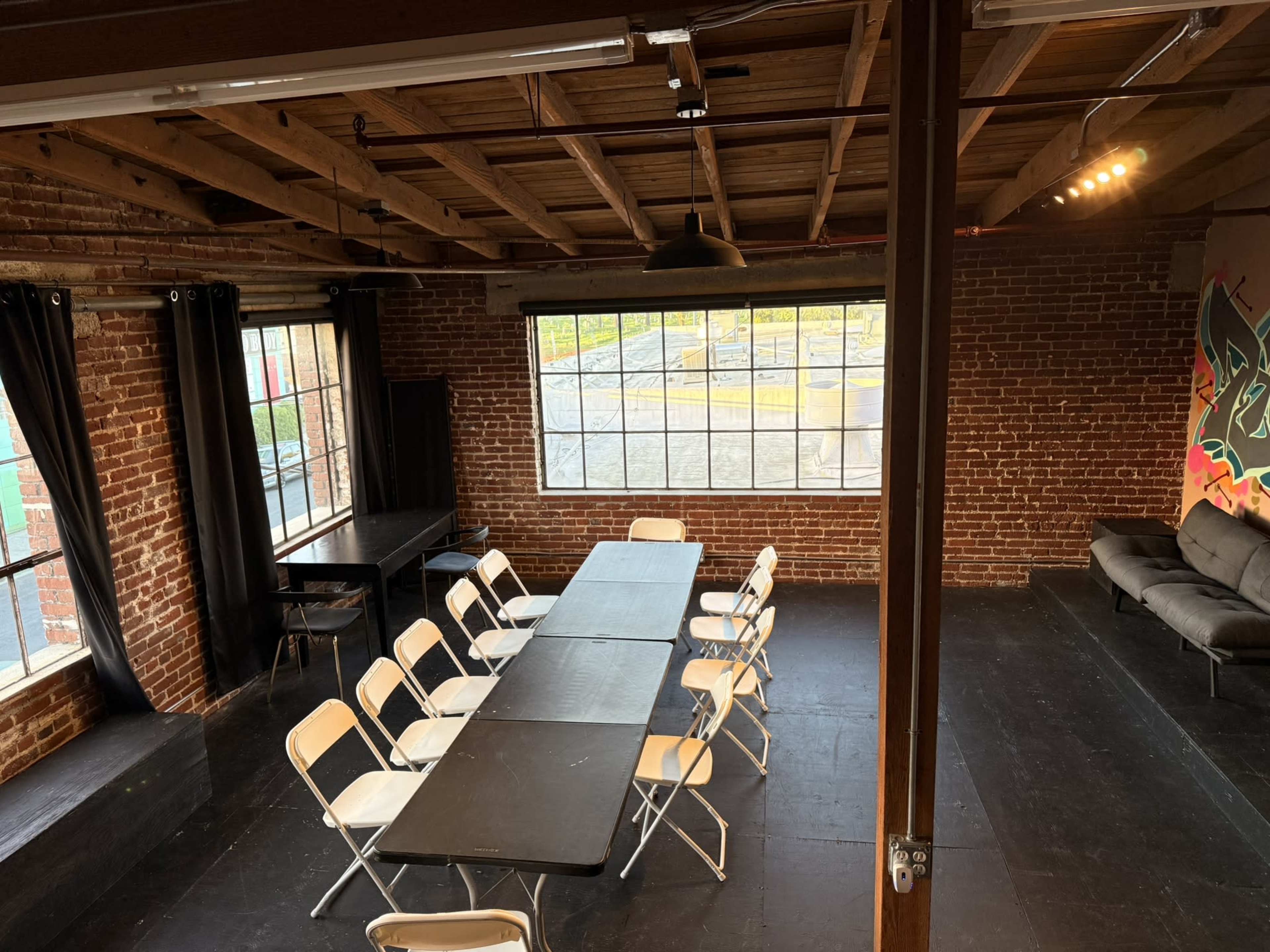 The image shows an industrial-style meeting room with exposed brick walls, a long black table, and white folding chairs arranged around it, illuminated by natural light through large windows.