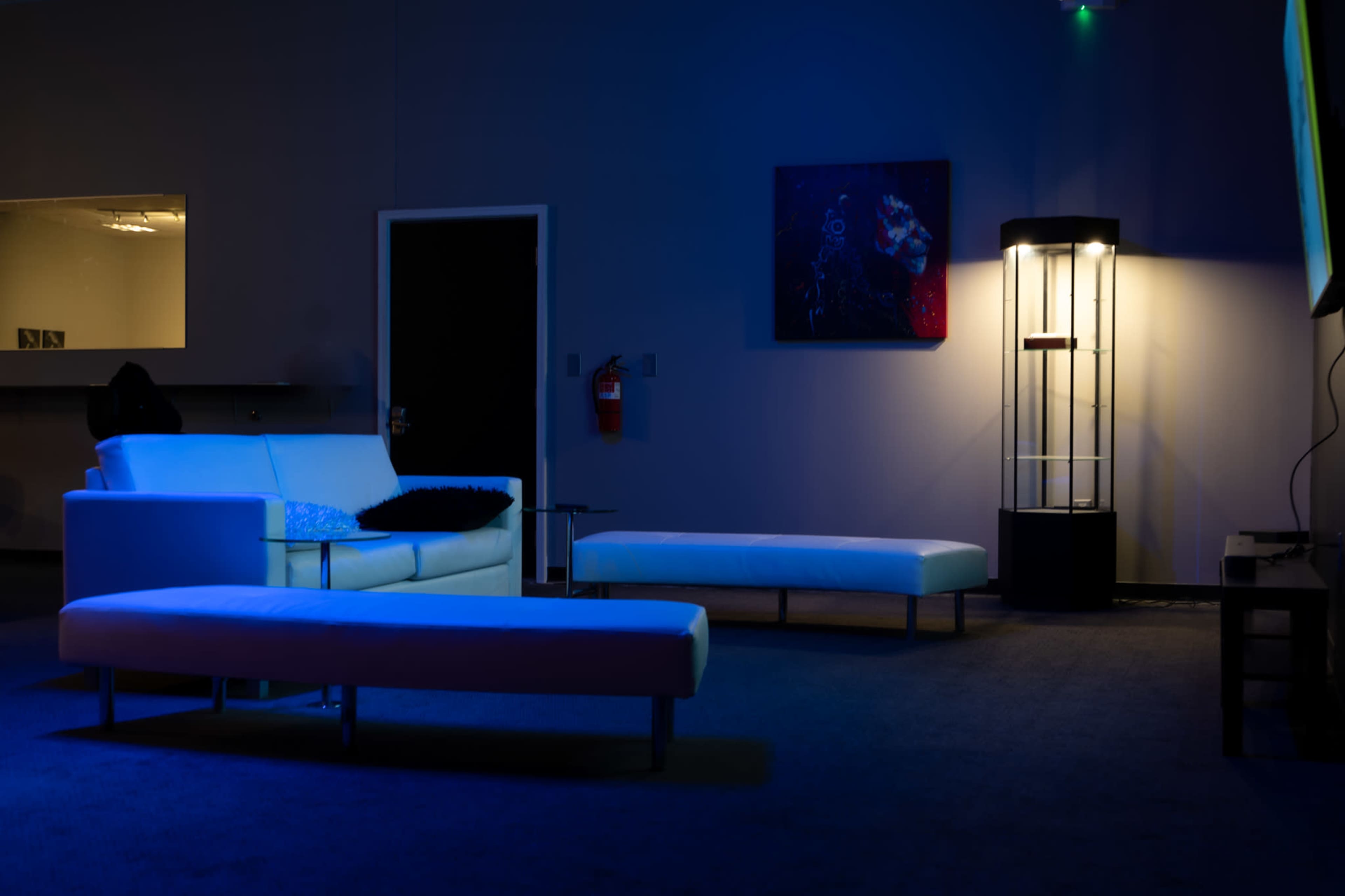 The room features a white sofa and two white benches under blue lighting, with a black-and-white wall art piece and a lit display case in the corner.