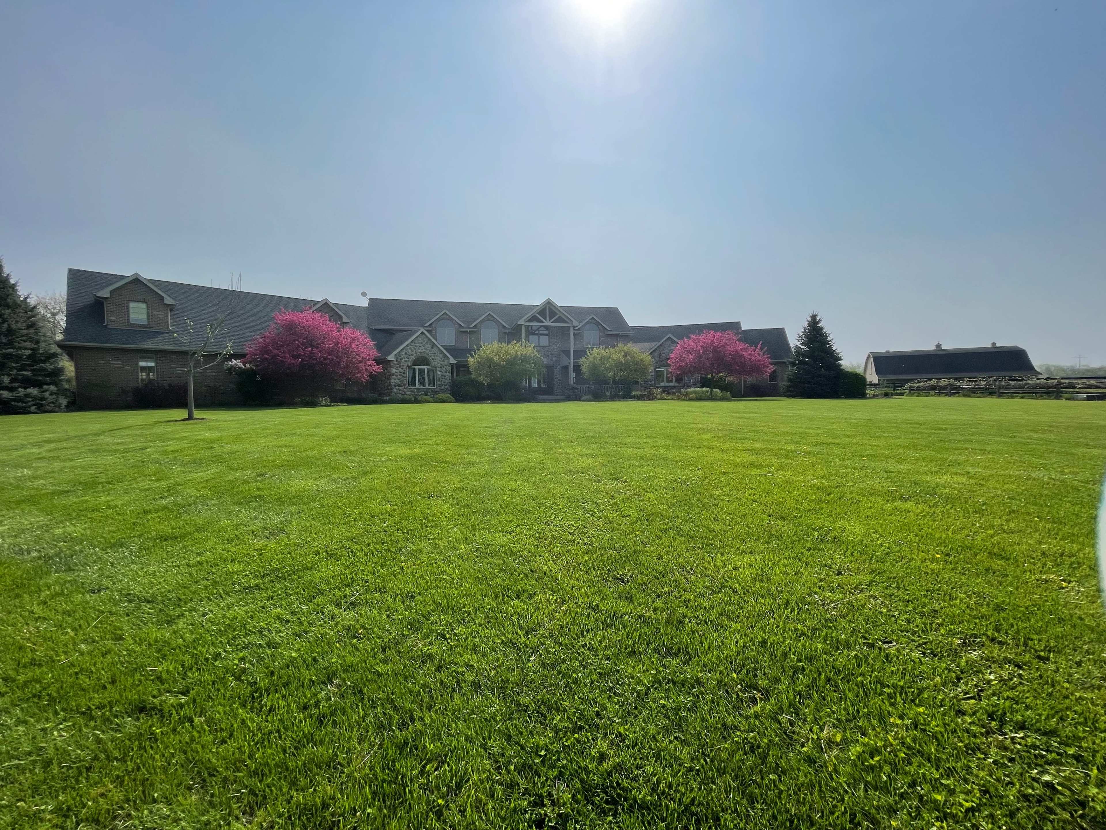 A large, two-story house with landscaped gardens and pink flowering trees sits on a vast green lawn.