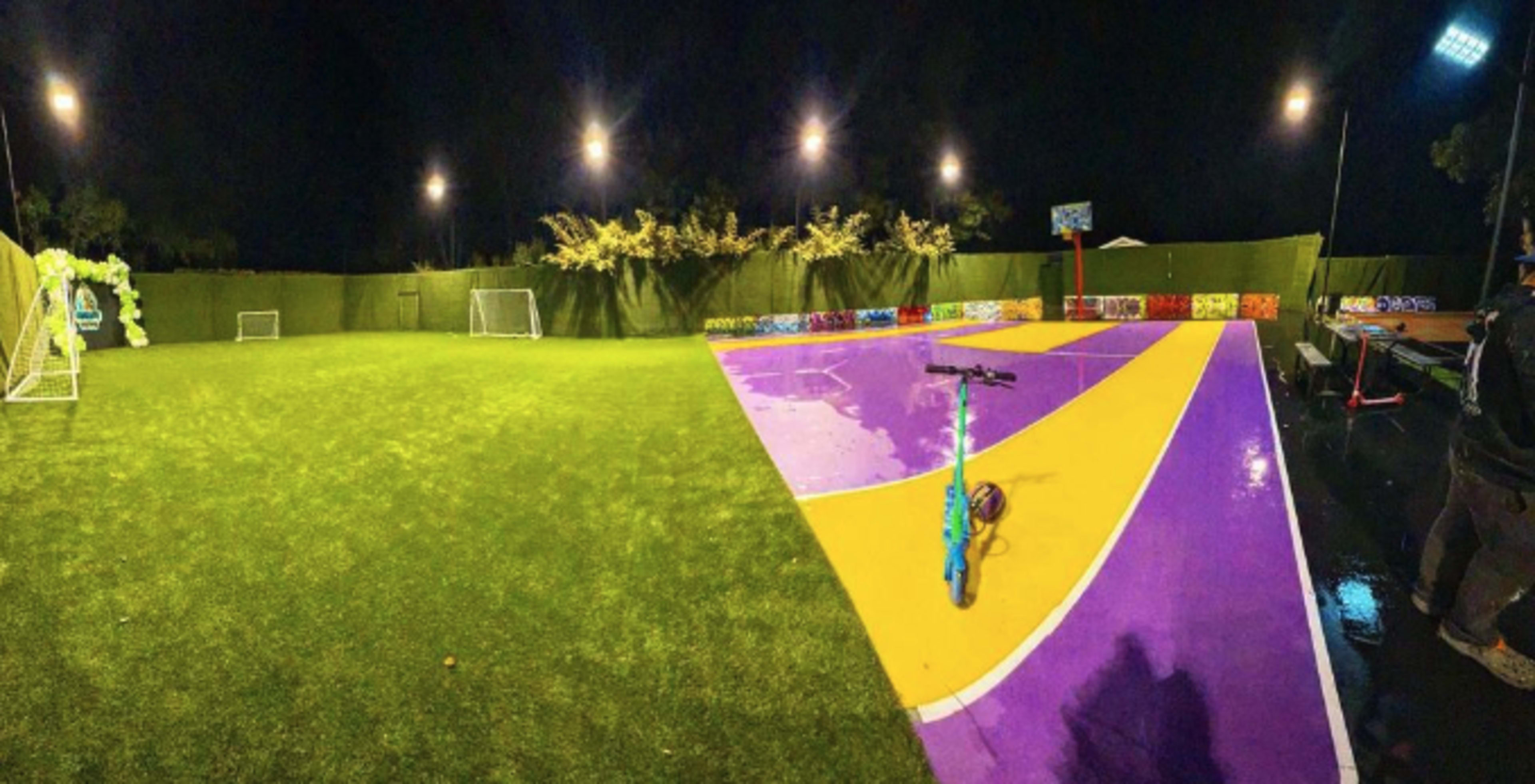 A brightly lit recreational area featuring a divided turf field with soccer goals on one side and a colorful, wet court on the other.