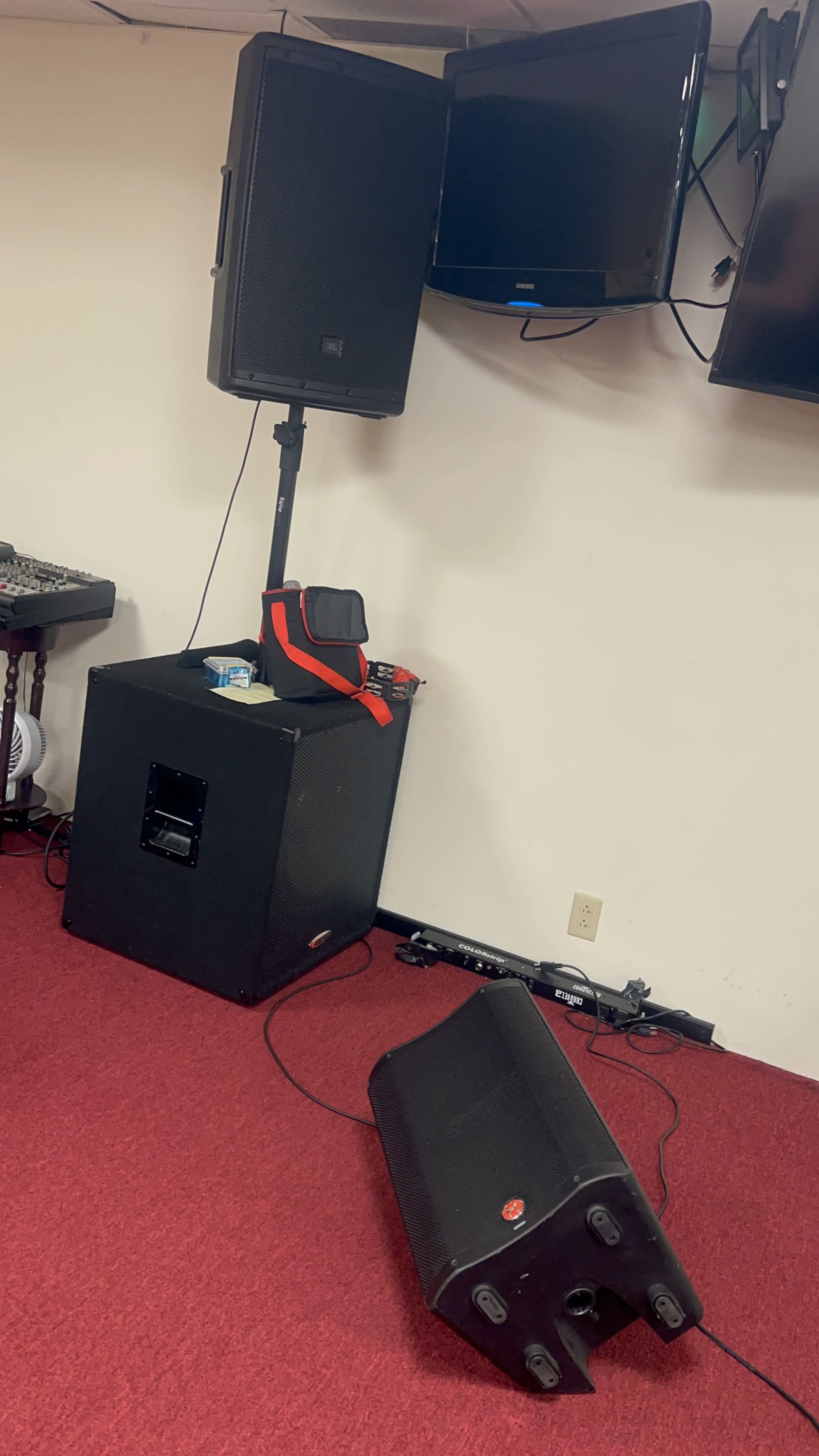 The image shows a room with a red carpet, featuring a speaker system, a soundboard, and two mounted televisions on the wall.