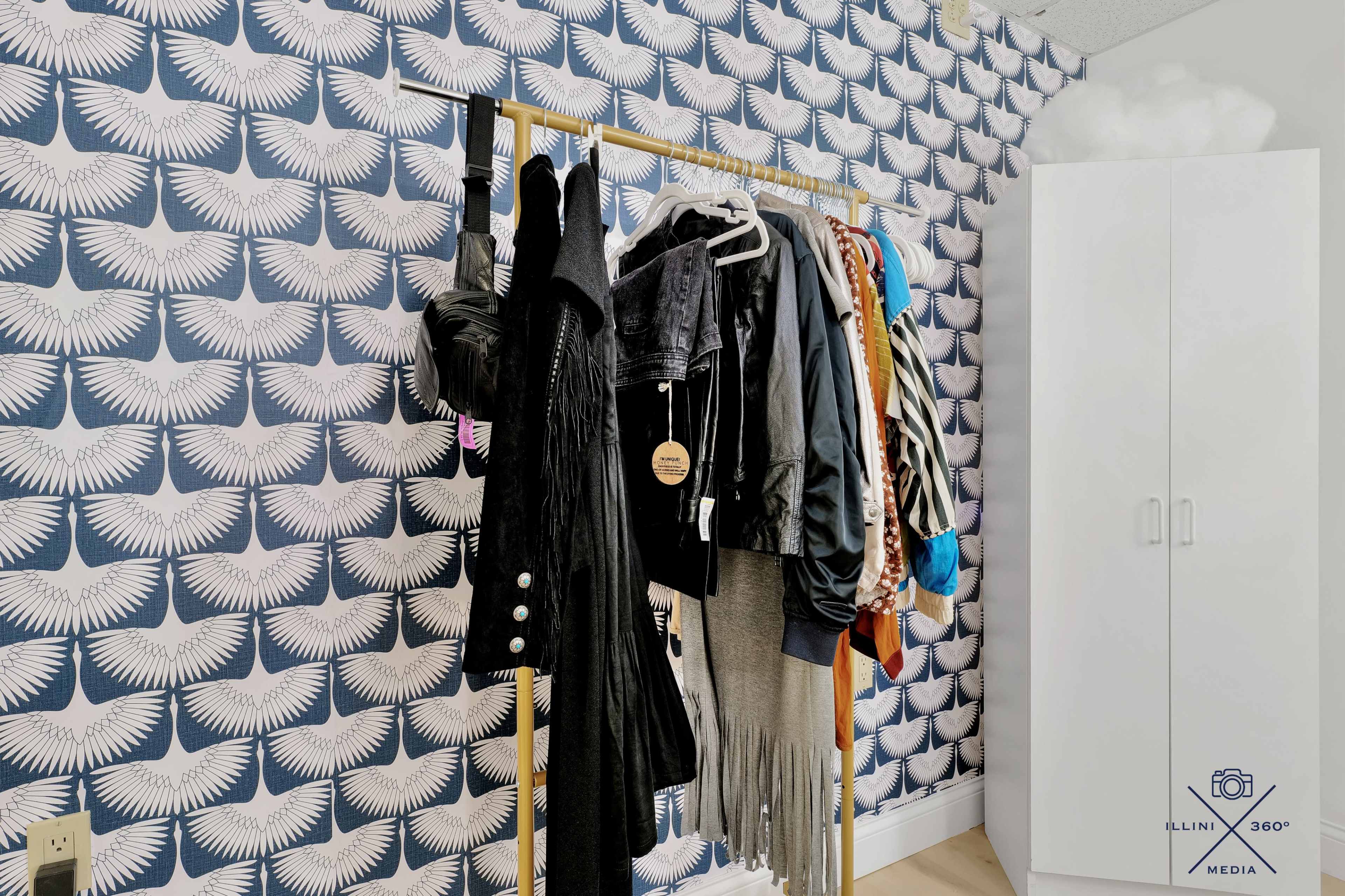 The image shows a clothing rack with various garments hanging against a patterned wallpaper featuring white feathers, alongside a white storage cabinet.