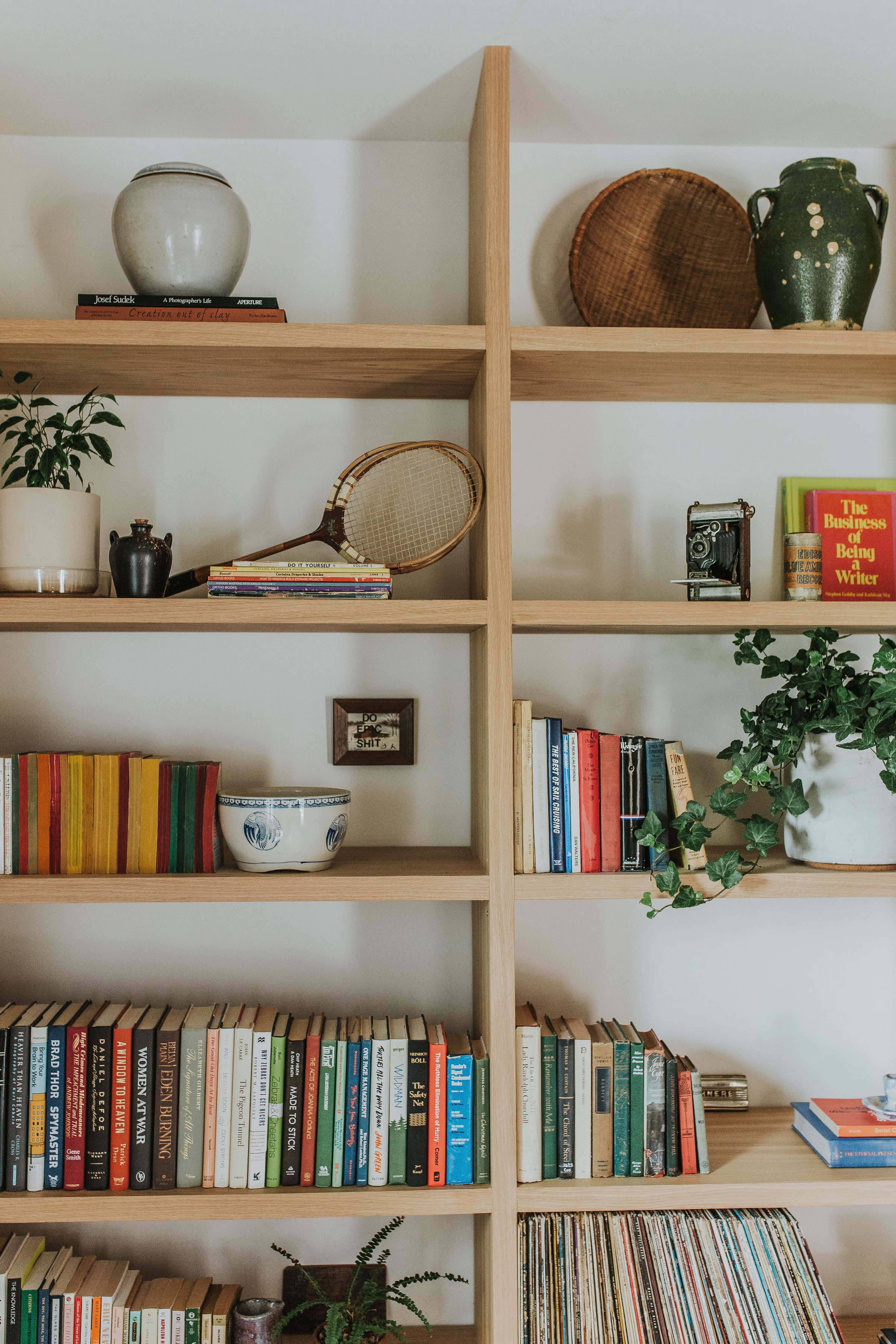 A wooden bookshelf displays a variety of books, plants, and decorative items, including a vintage camera and a tennis racket.