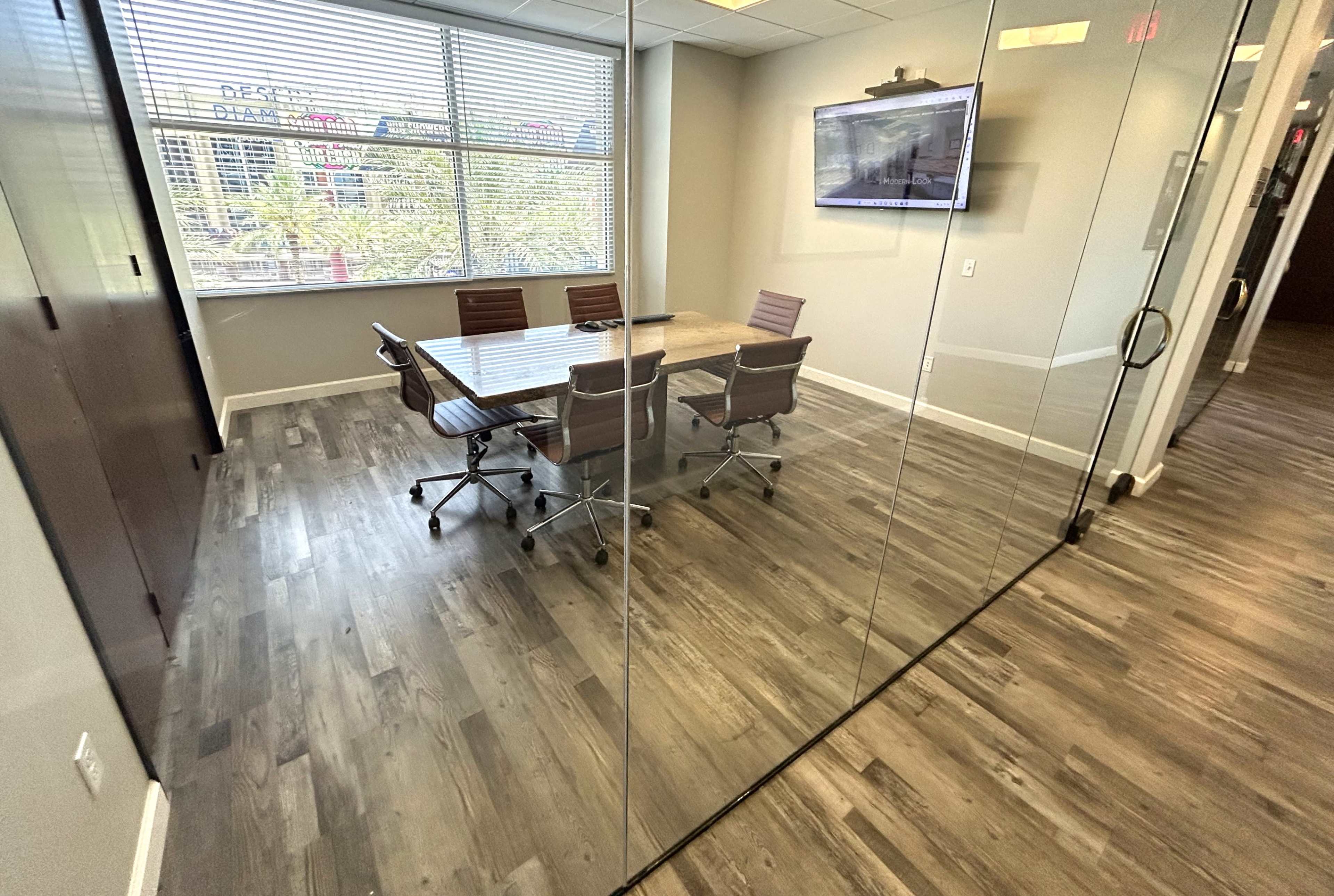 A modern conference room features a wooden table surrounded by brown leather chairs, with large windows offering a view of the outdoors.