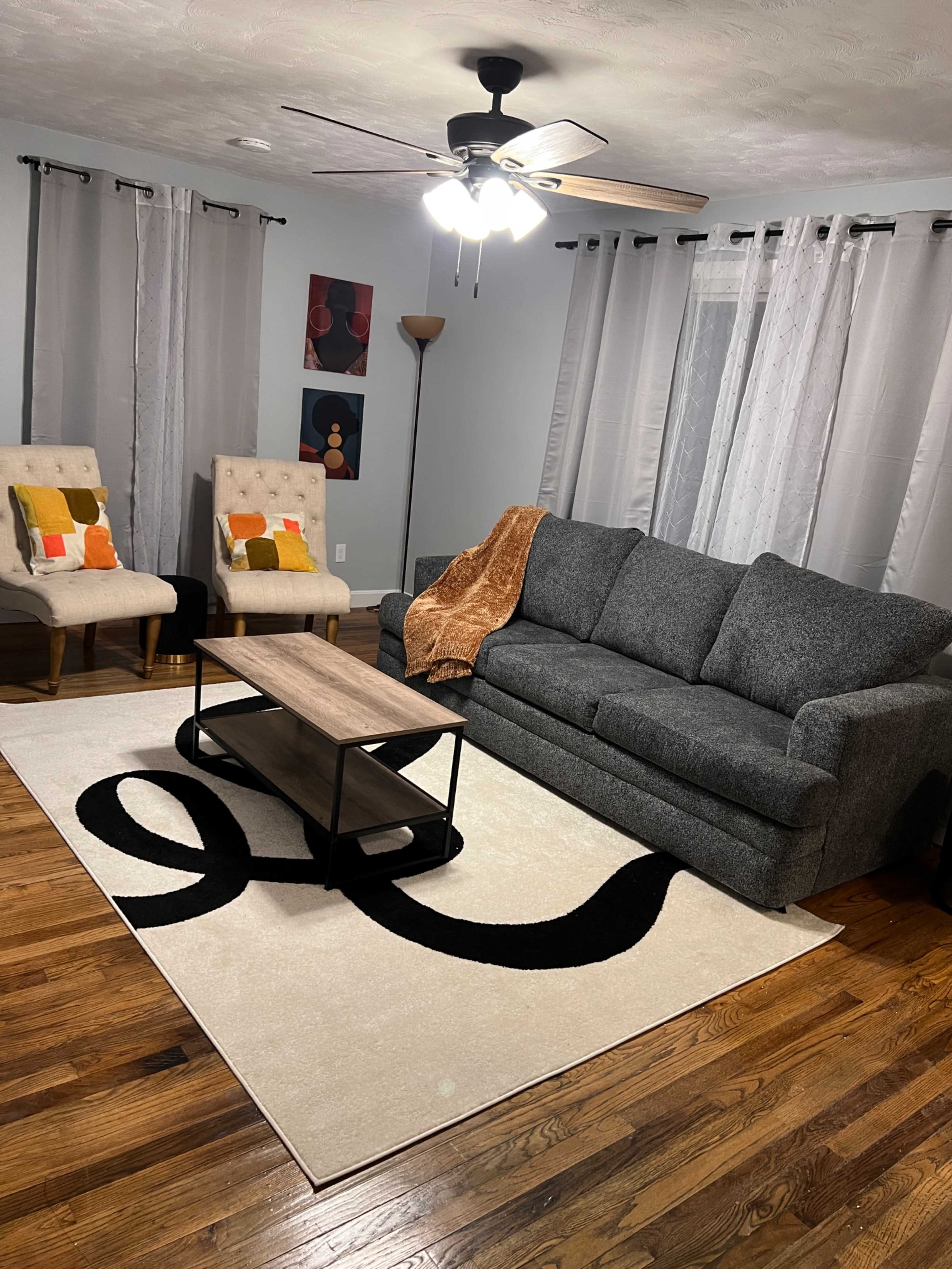 The living room features a gray sofa, two upholstered chairs, a wooden coffee table, and a patterned area rug, all arranged on hardwood flooring.