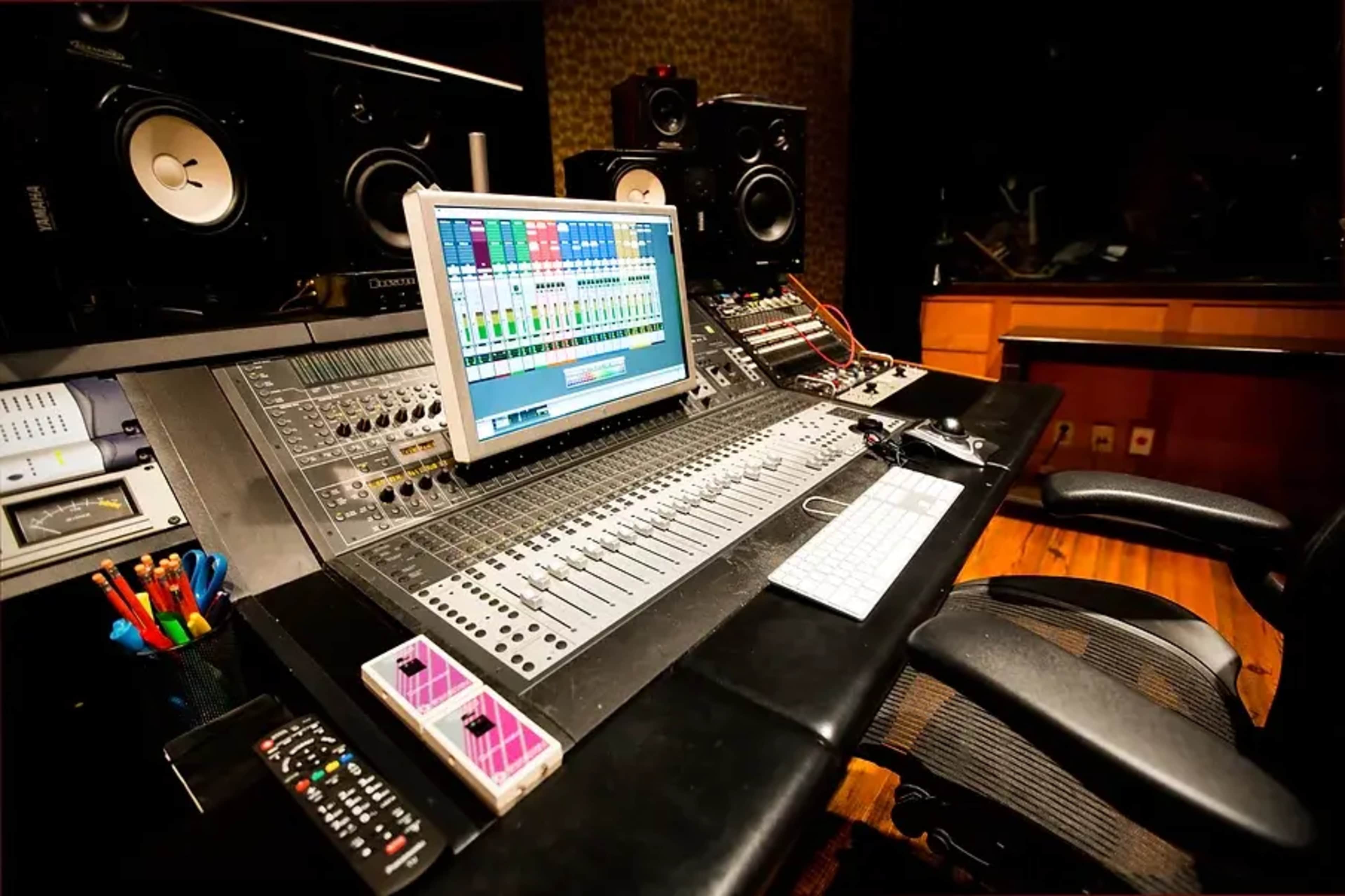 A music production studio with a digital mixing console, computer monitor, and speakers arranged on a workstation.