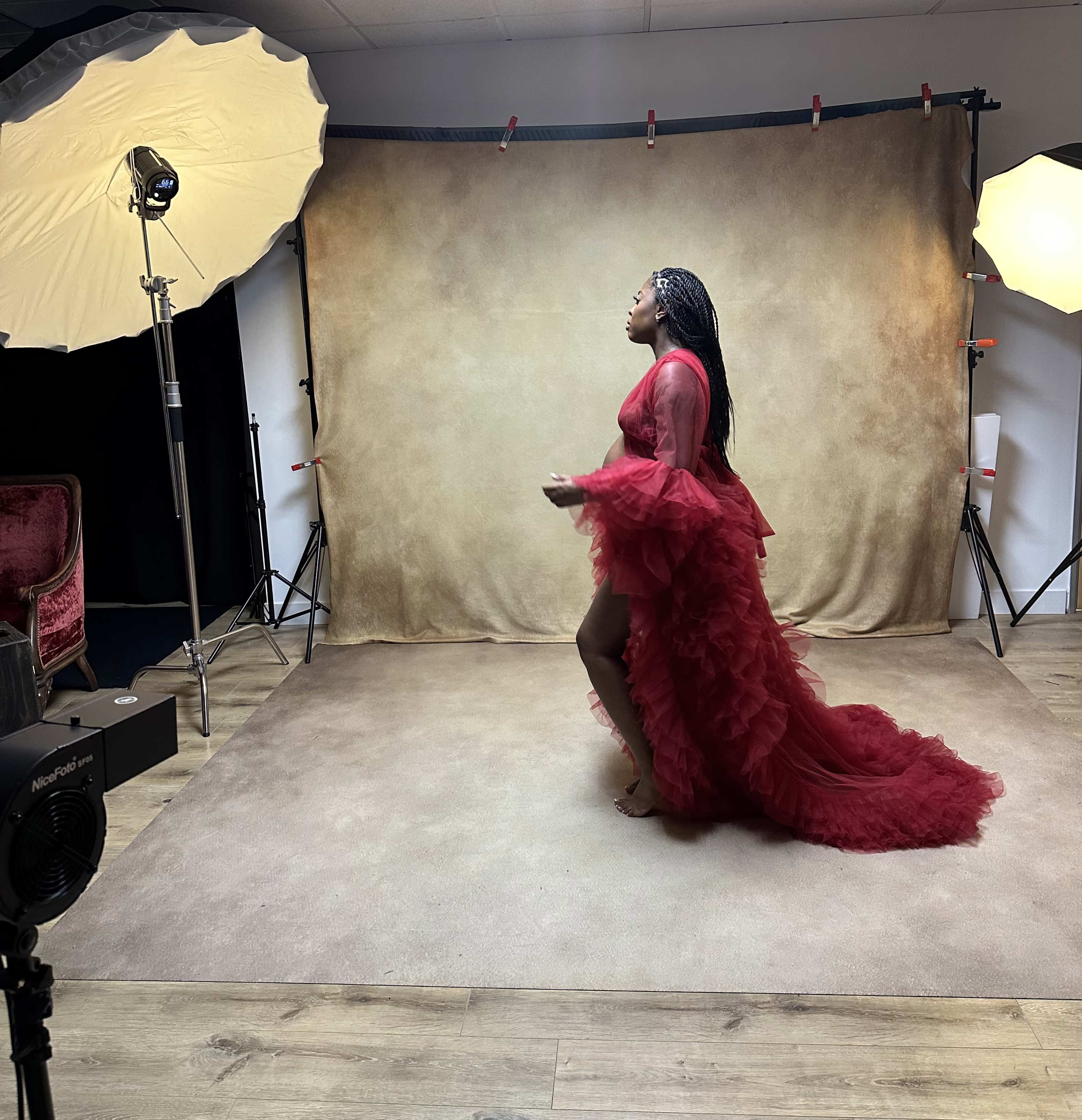 A model in a red dress poses on a beige backdrop in a photography studio.