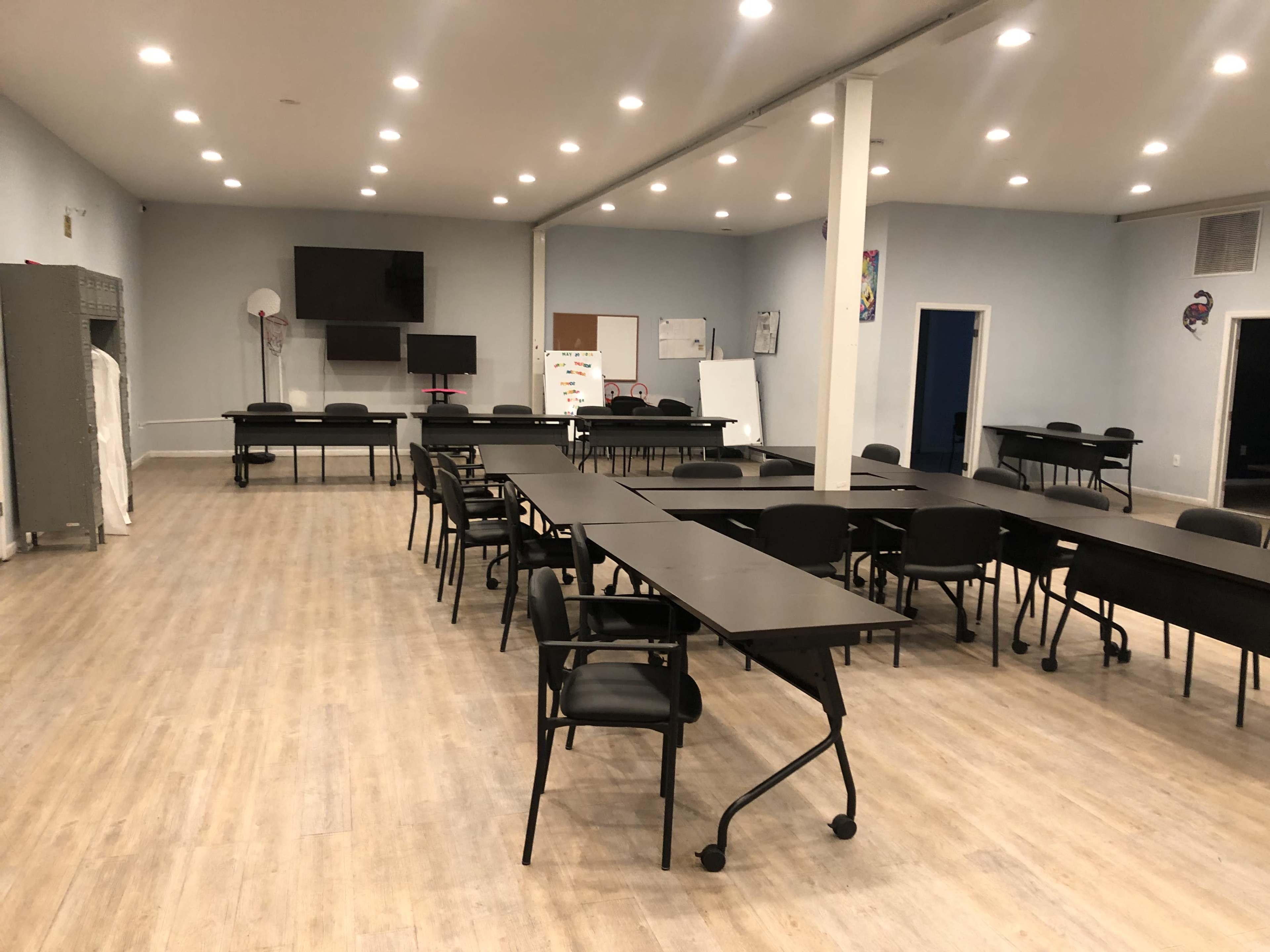 The image shows a spacious, empty classroom setup with several long black tables and chairs arranged in a professional layout, illuminated by bright ceiling lights.