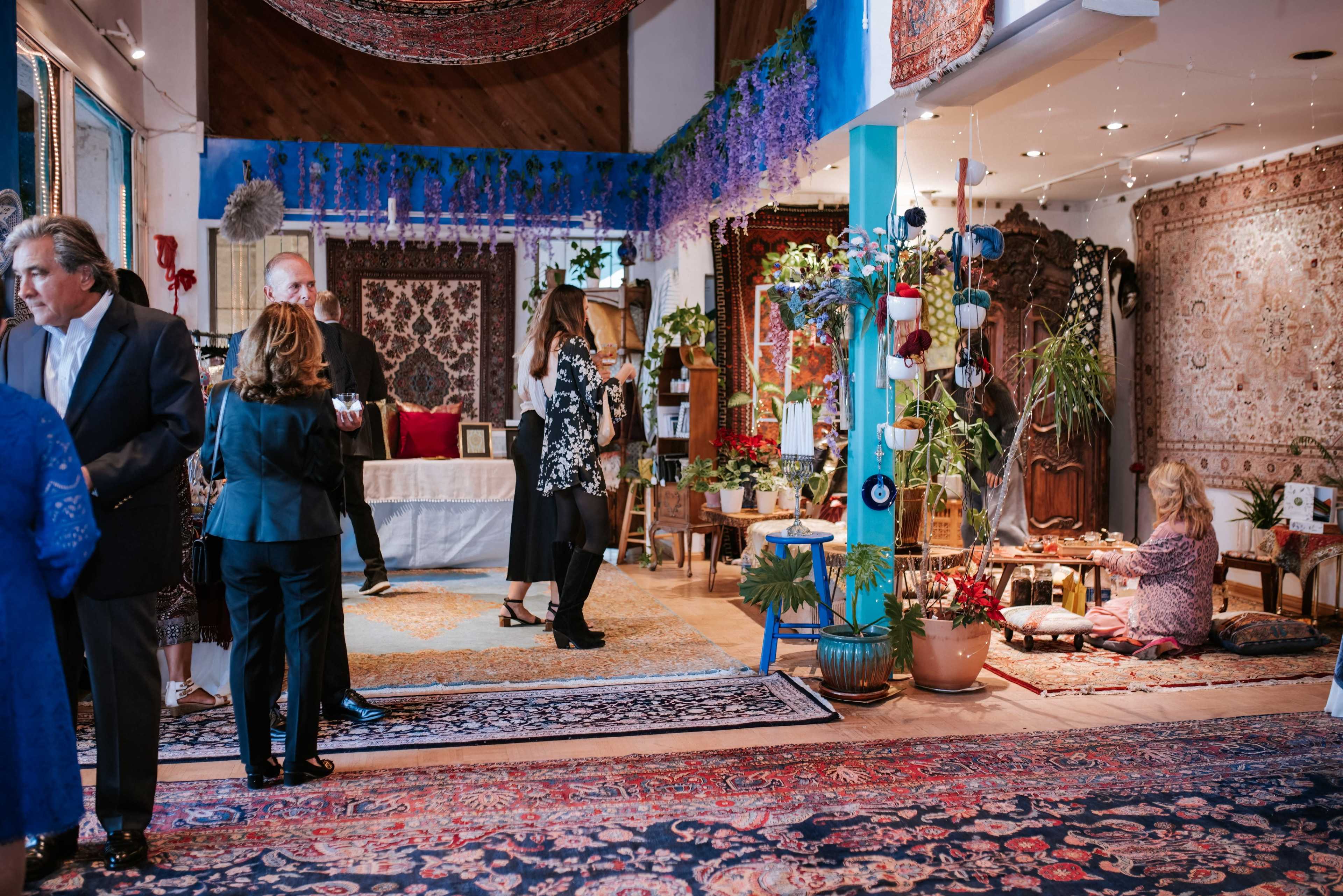 A group of people mingle in a warmly decorated space filled with intricate rugs, plants, and decorative tapestries.