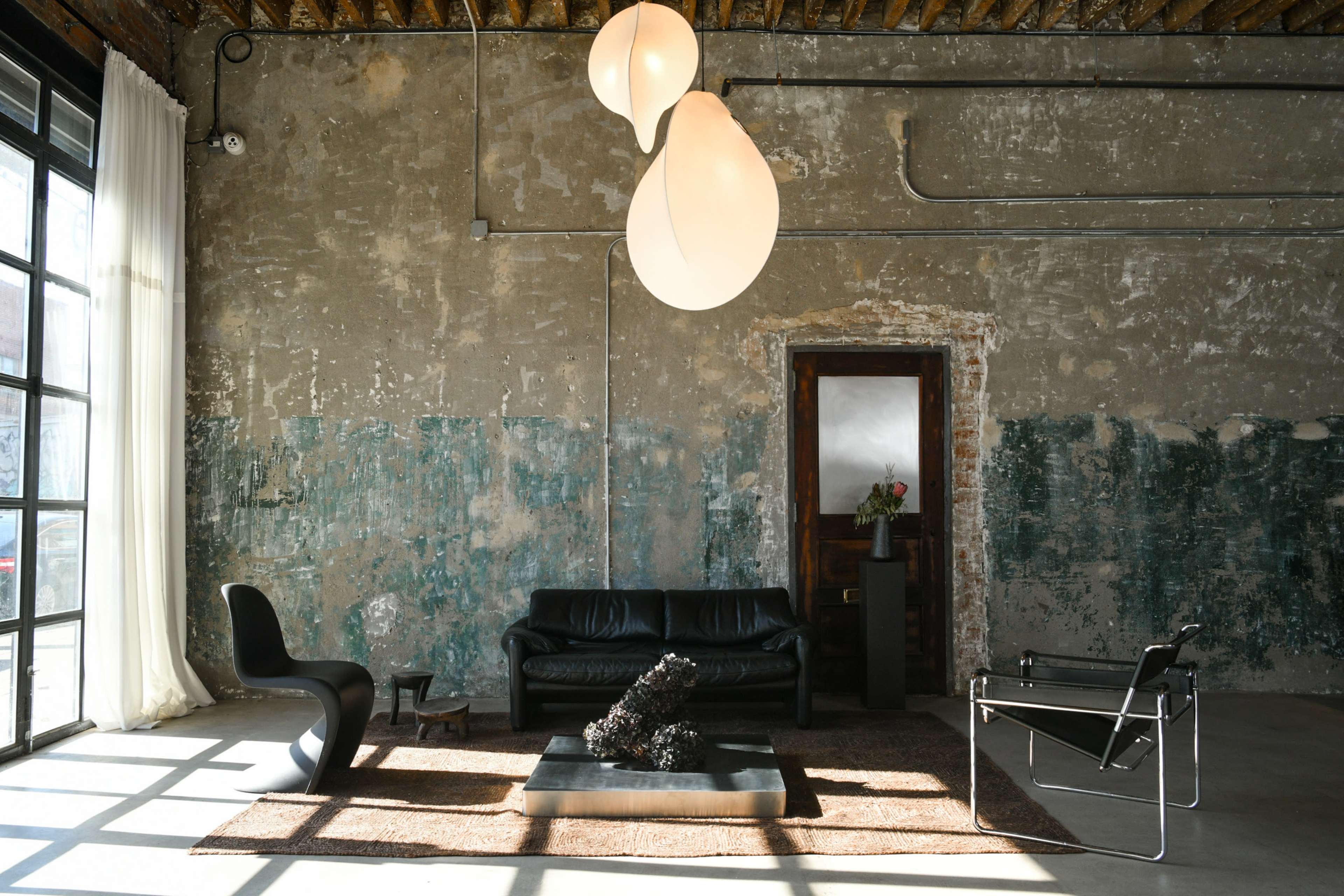 The image shows a minimalist interior with a black leather sofa, a unique lighting fixture, and a coffee table placed on a textured rug, set against a weathered wall.