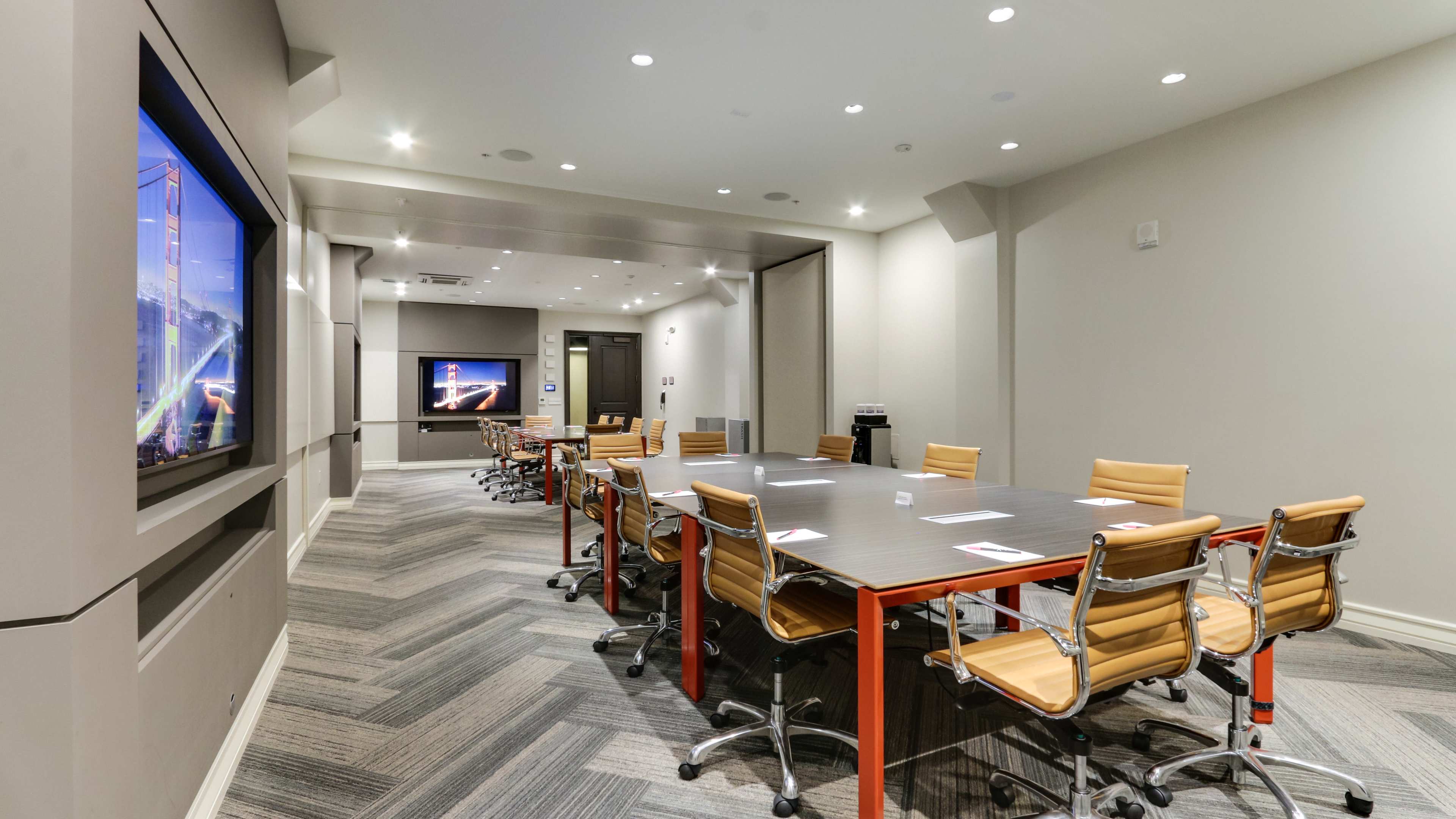 The image shows a modern conference room with a long rectangular table surrounded by chairs and two large screens on the walls.