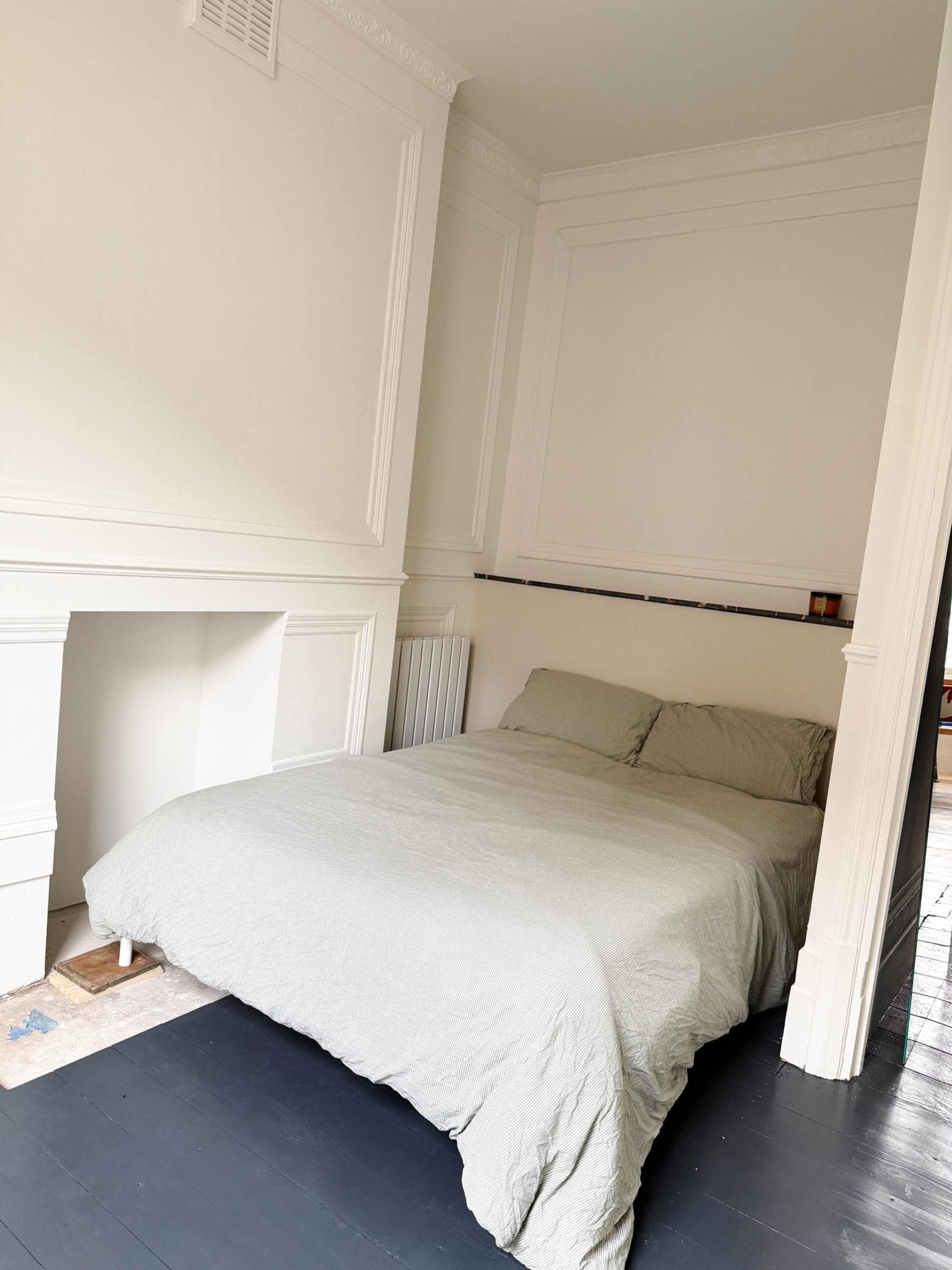 The image shows a small bedroom with a bed covered in a light grey blanket, positioned against a wall adorned with minimalist moldings.
